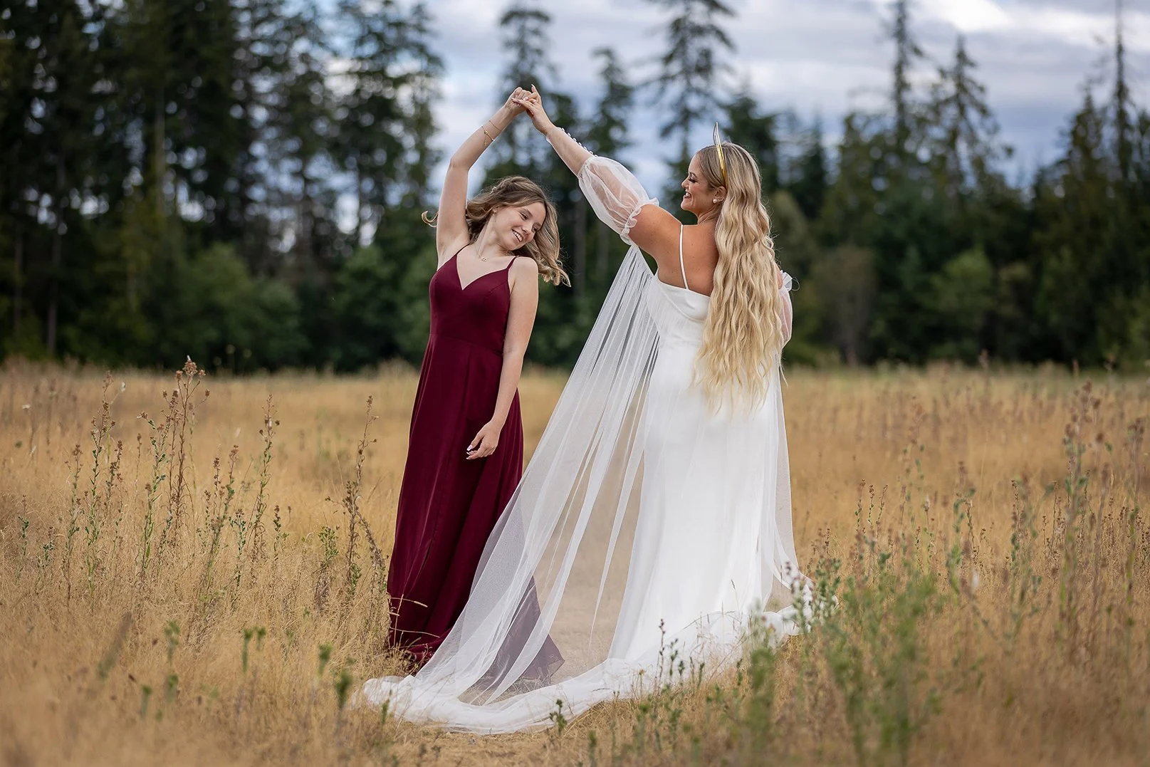 Two women, one in a white flowing dress with sheer, puffy sleeves and a long train, and the other in a sleeveless burgundy dress, dancing and holding hands in a grassy field with trees in the background.