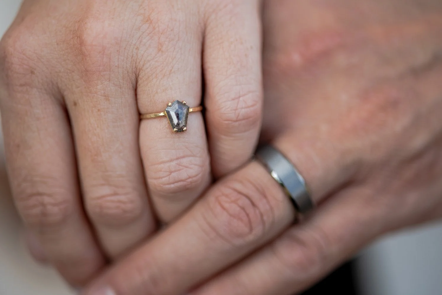 Close-up of a woman's hand with a gold ring featuring a large, raw, irregular-shaped gemstone, and a man’s hand with a simple silver band, both wearing wedding rings.