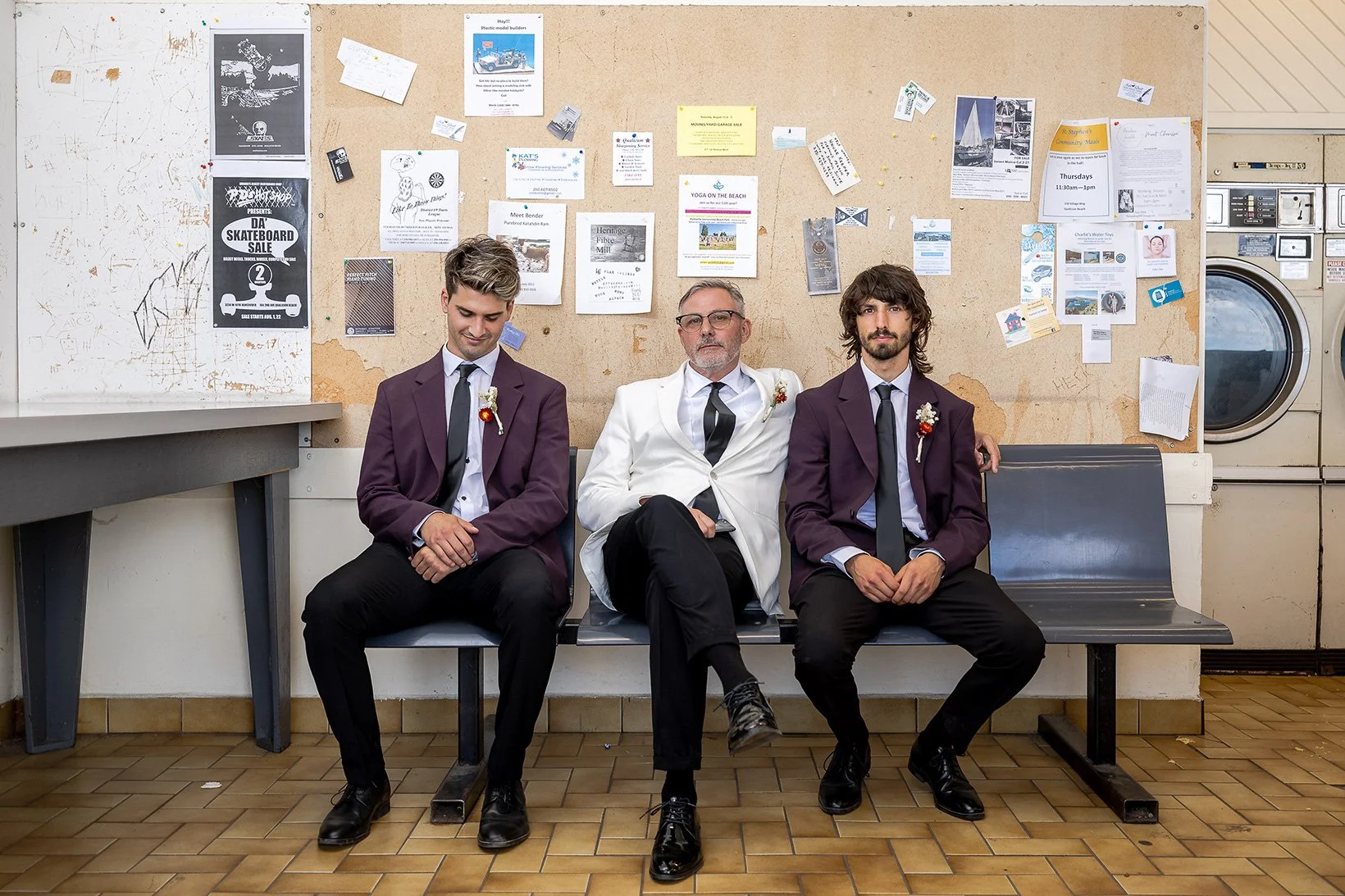 Three men sitting on a bench inside a laundromat. The man in the center is wearing a white suit, and the other two men are wearing dark purple suits. The background has a bulletin board with various notices and a washing machine.