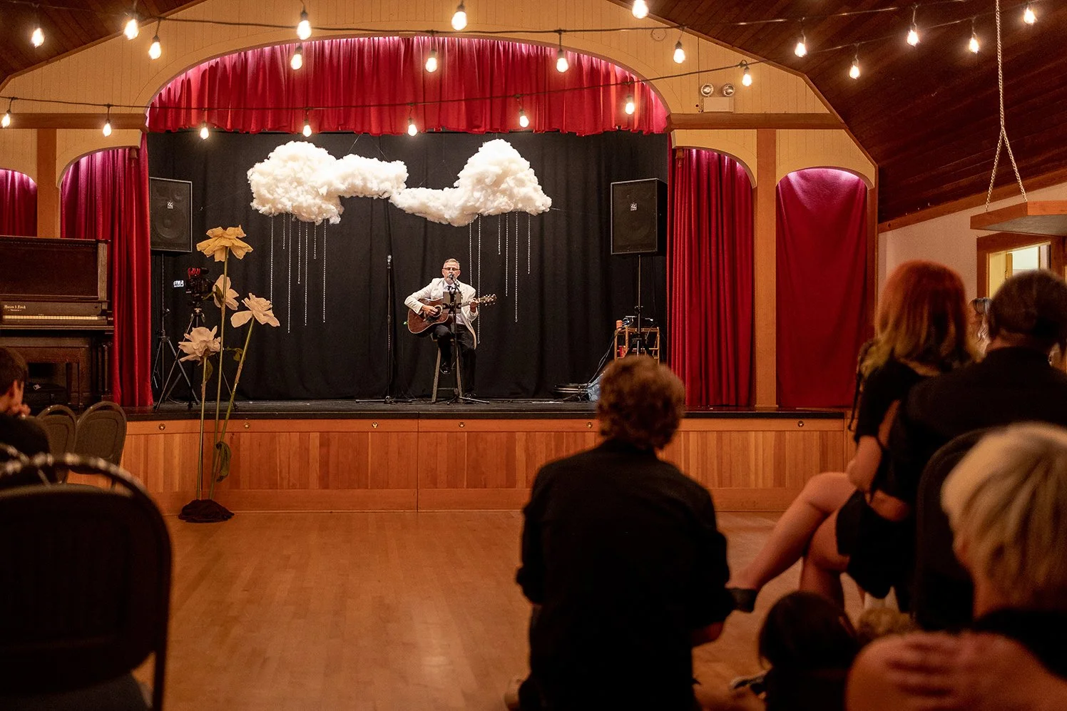 Indoor stage with a man playing guitar and singing, decorated with hanging clouds and flowers, red curtains and string lights, audience seated in front.