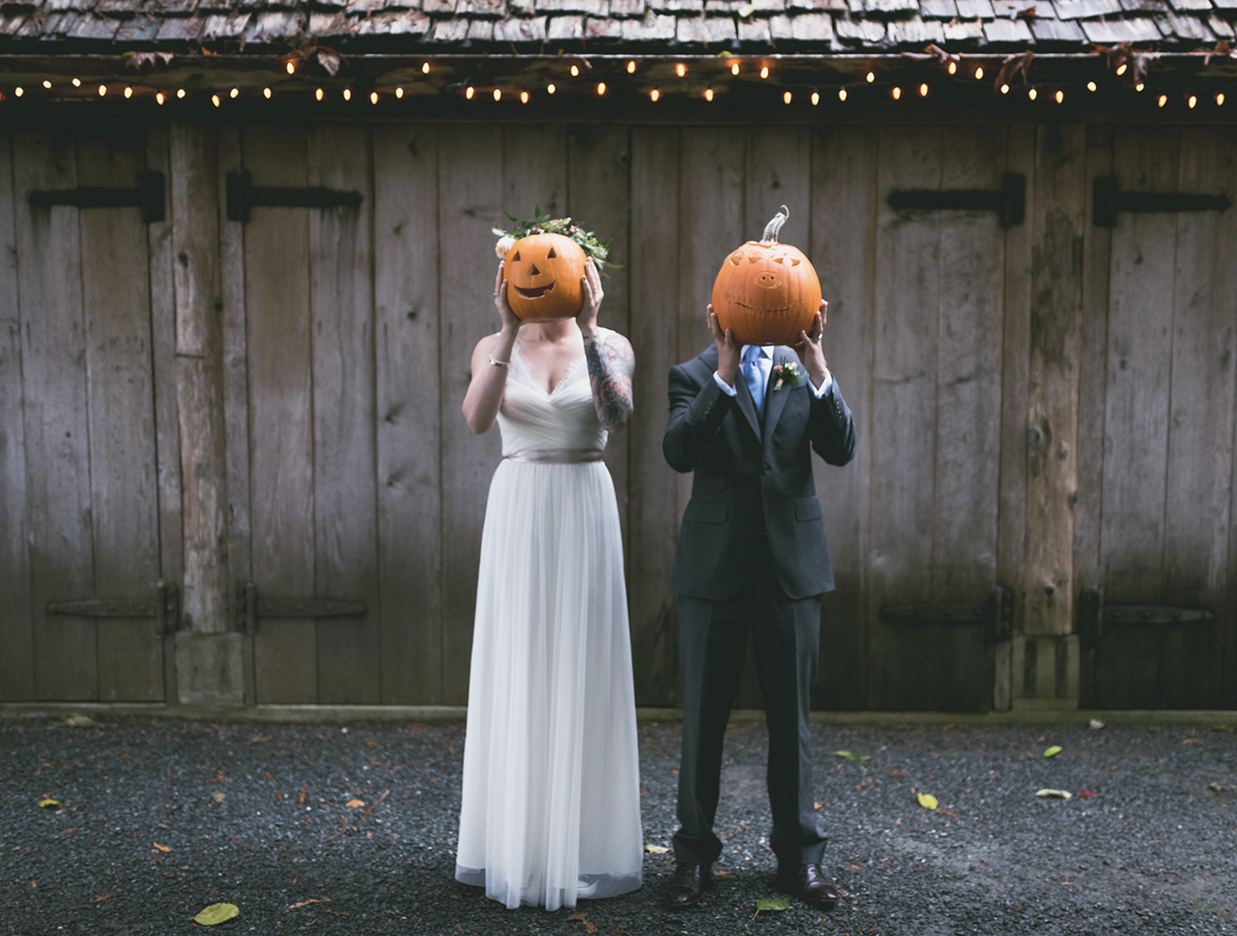 A bride and groom standing in front of a rustic wooden fence, each holding a carved pumpkin mask in front of their faces. The bride is wearing a white wedding dress with tattoos on her left arm, and the groom is dressed in a black suit with a boutonniere. String lights are hanging above them, and there are fallen leaves on the gravel ground.