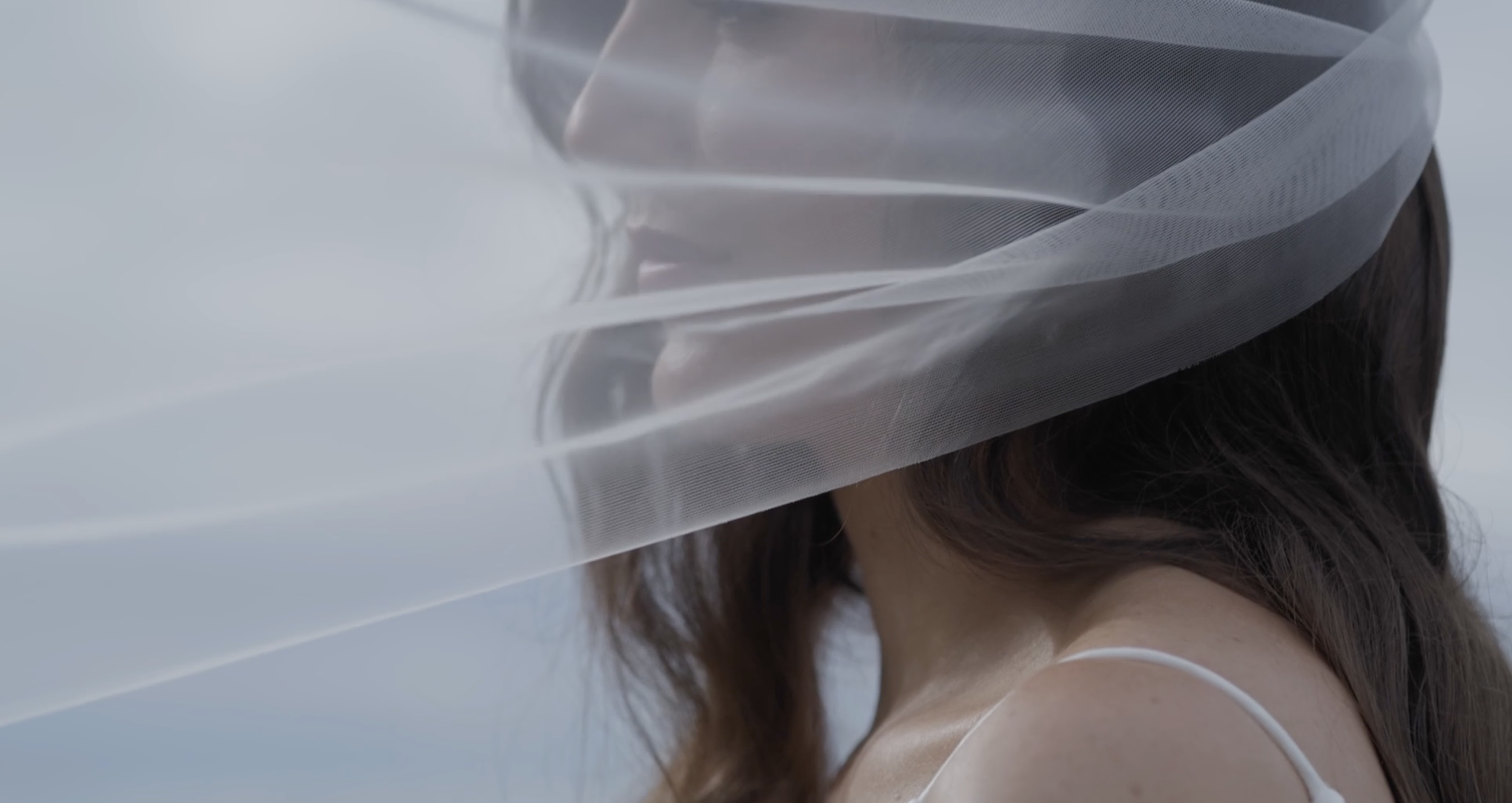 A person with long dark hair wearing a white spaghetti strap top, partially obscured by a sheer, light-colored fabric or veil.