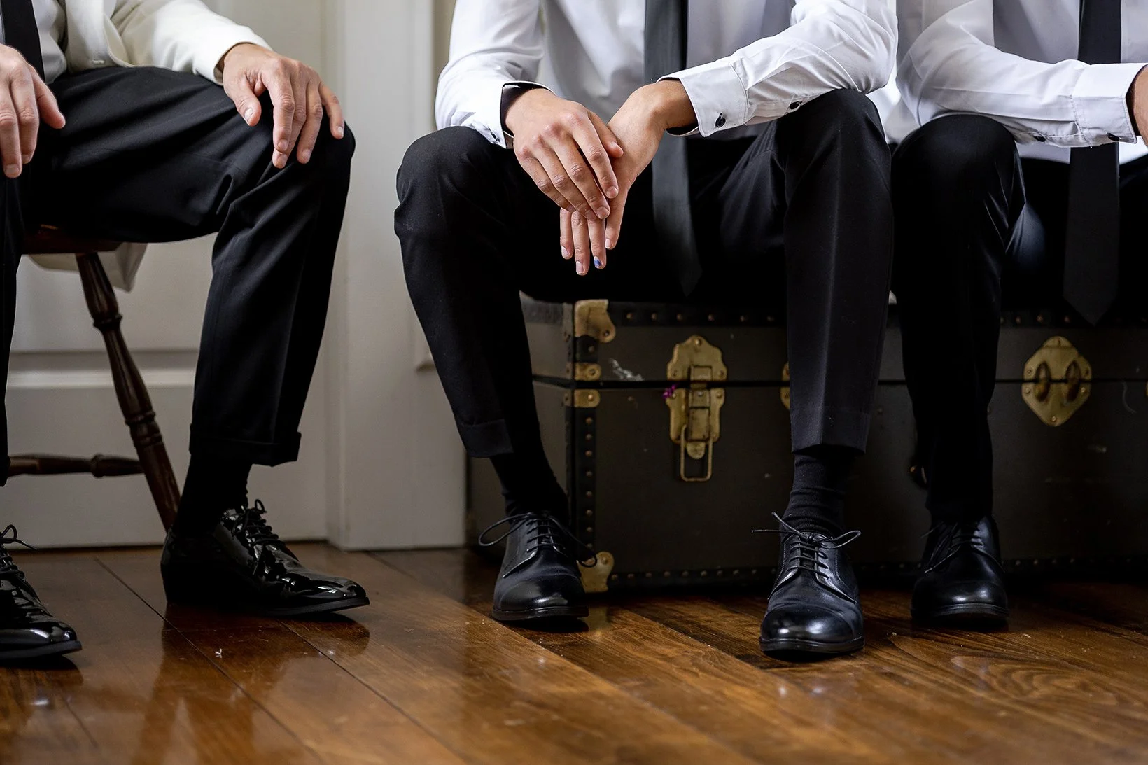 Three men sitting, wearing black dress shoes and black pants. Two of them are dressed in white shirts, and next to one man, he is holding hands with the other.