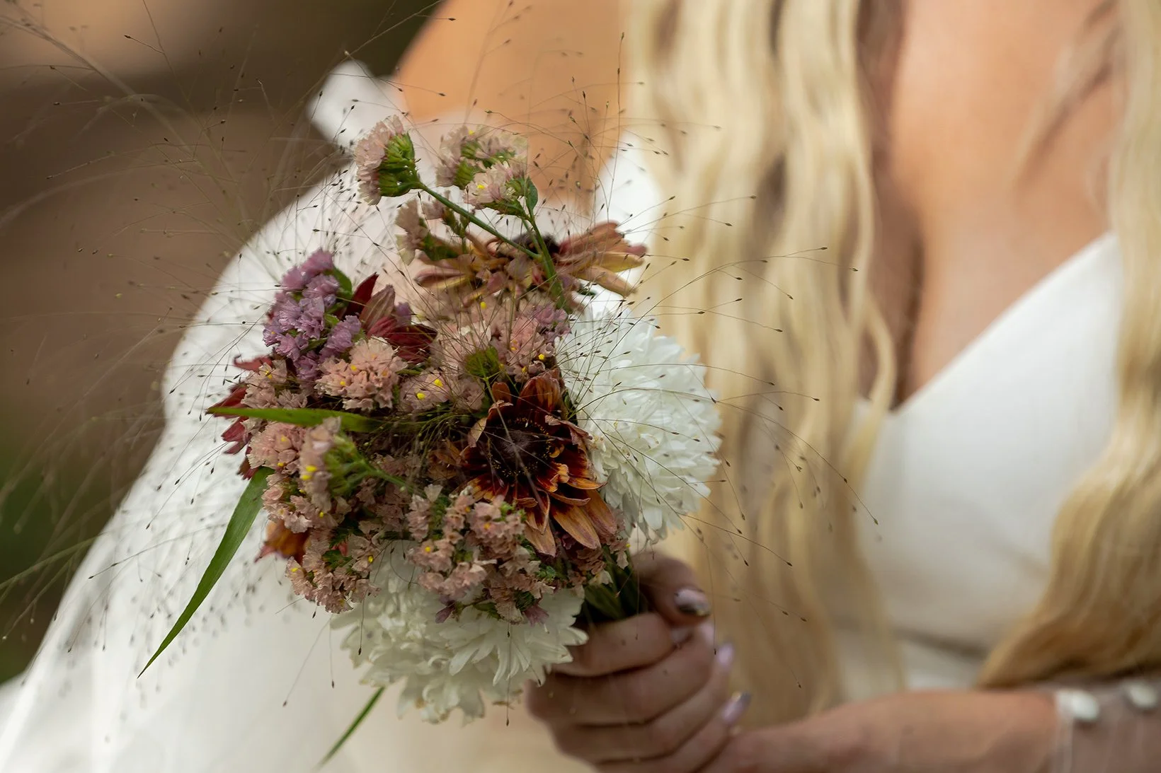 A woman in white holding a bouquet of pink, purple, red, and white flowers with green stems and delicate wispy grass accents, close-up of her face partially obscured, with long blonde hair and wearing a face mask.