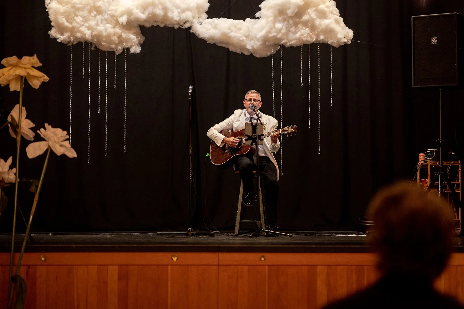 A man in a cream-colored blazer and glasses playing an acoustic guitar on a stage decorated with a large cloud made of cotton and hanging chains, with flowers on the left side and a speaker on the right side.