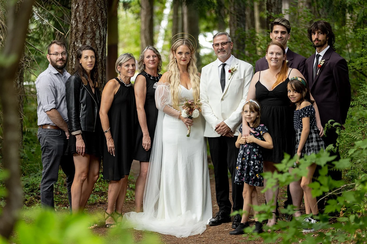 A wedding celebration in a wooded outdoor area with multiple family members, including a bride in a white gown holding a bouquet, a groom in a white suit, and children dressed in black and polka dots.