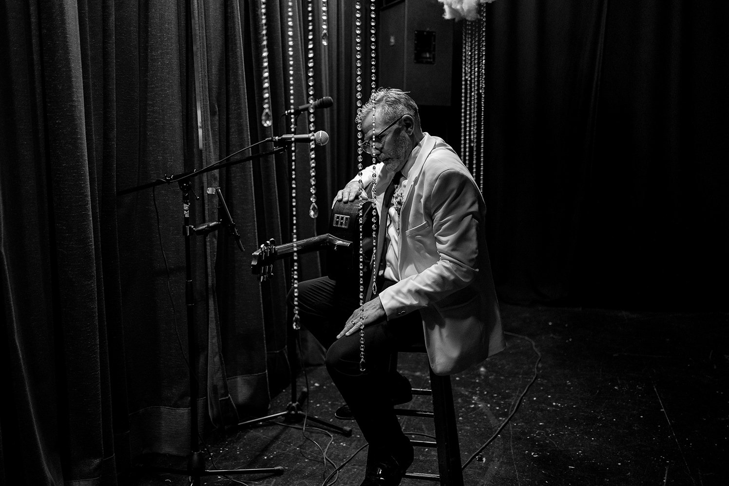 Black and white photo of a man dressed in a light-colored blazer sitting on a chair on stage, holding a guitar, with a microphone in front of him and stage curtains in the background.