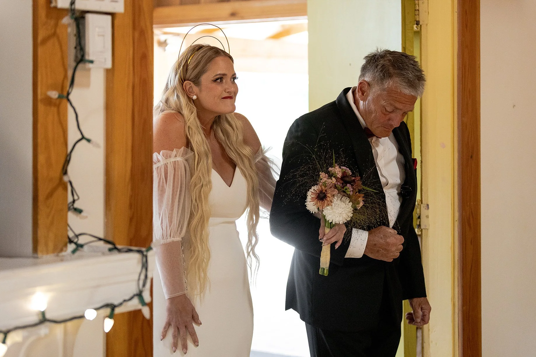 A woman in a white dress with long blonde hair looking thoughtfully, and a man in a black tuxedo holding a bouquet of flowers, standing in a doorway with wooden trim and string lights.