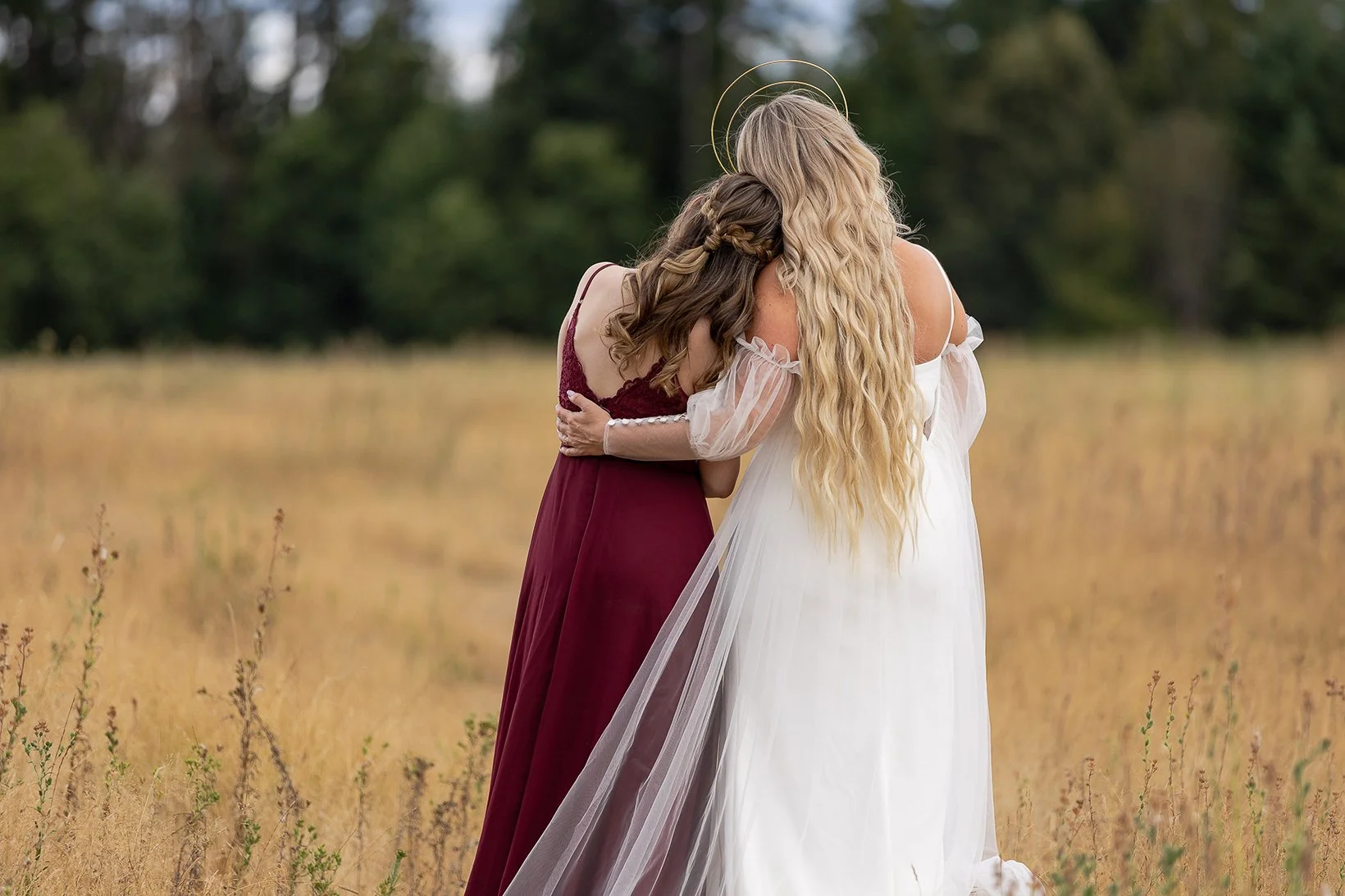Two women in wedding attire embracing in a field of tall grass with trees in the background.