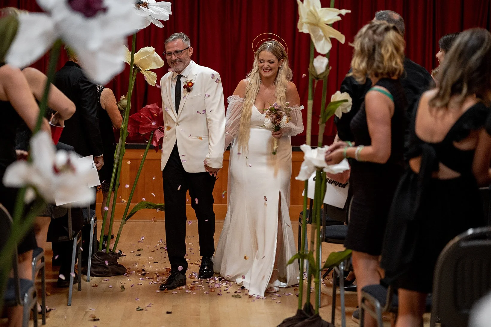 A newly married couple walks down the aisle holding hands, smiling, surrounded by guests on both sides, with flower petals on the floor, in a room with red curtains in the background.