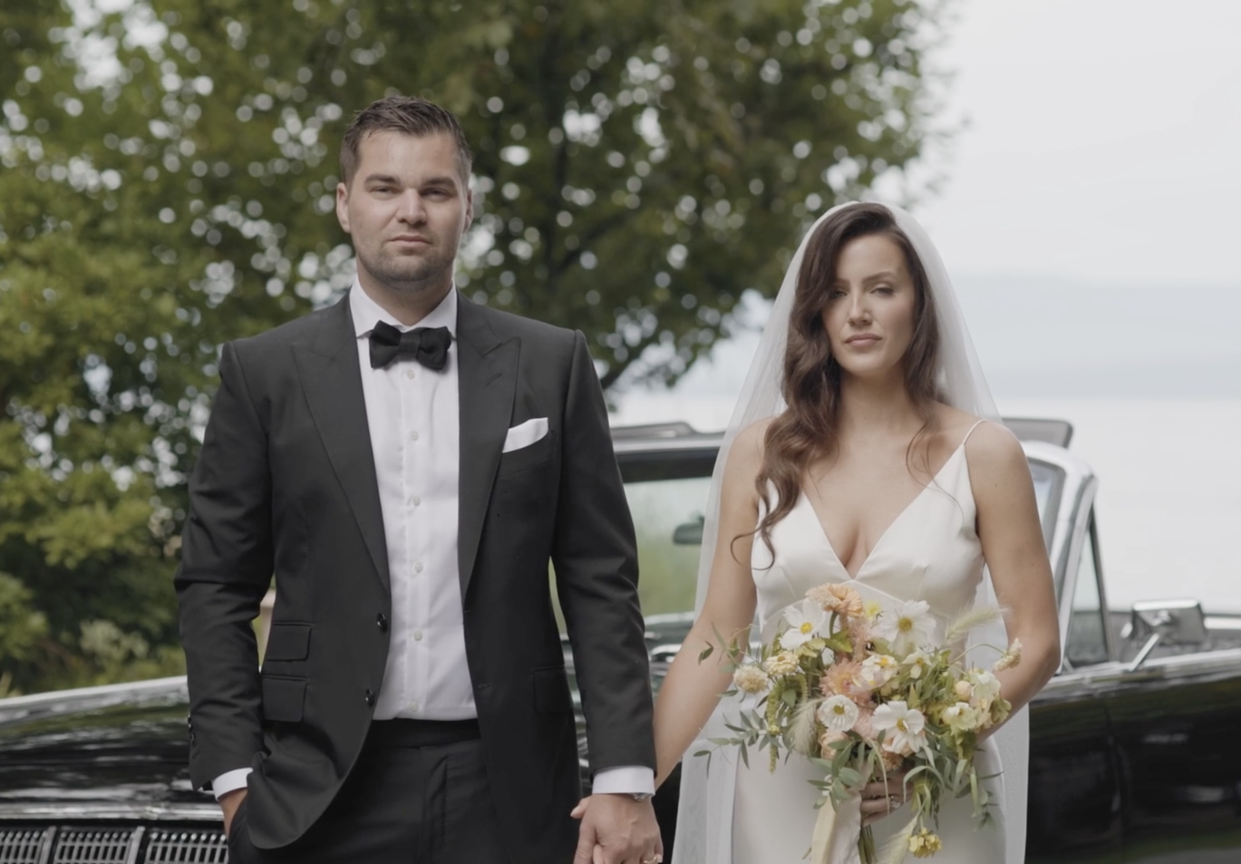 A newlywed couple standing outdoors in front of a black car, with trees in the background. The groom, in a black tuxedo and bow tie, has a serious expression. The bride, wearing a white wedding dress and veil, holds a bouquet of flowers and has a neutral expression.
