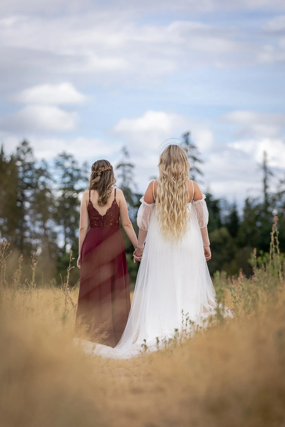 Two women holding hands walking in a field, one wearing a white dress with long blonde hair, the other in a dark red dress with brown hair, with trees and a blue cloudy sky in the background.
