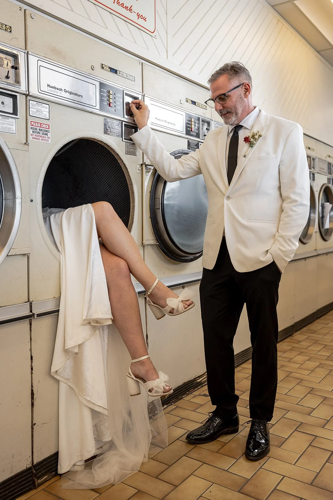 A person is inside a laundromat washing machine with their legs hanging out, wearing a white dress. A man in a white blazer, black pants, and shiny black shoes stands nearby, smiling and looking at the person in the washing machine.