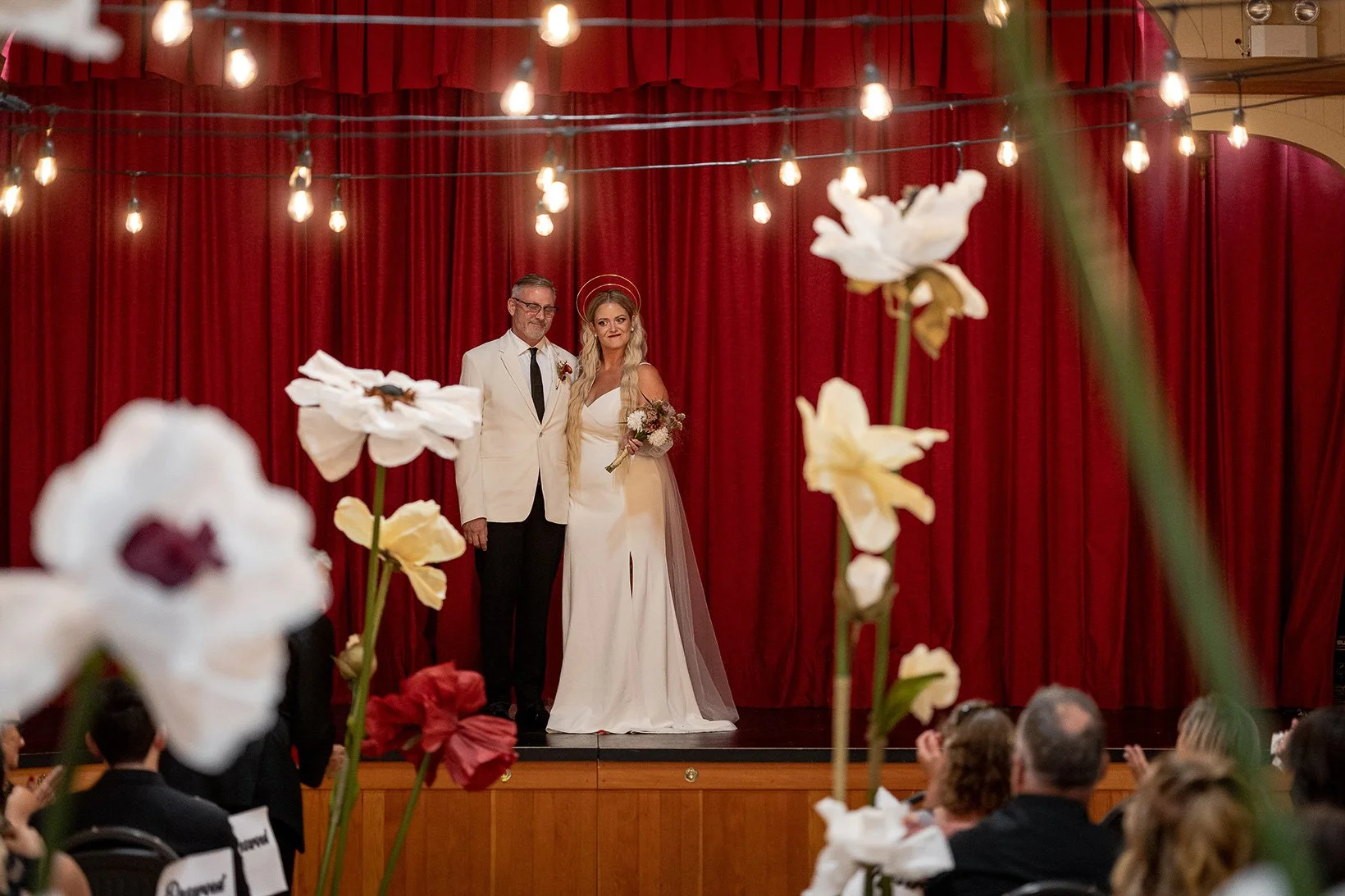 A bride and groom standing on a stage with red curtains, surrounded by flowers and string lights, at a wedding celebration.