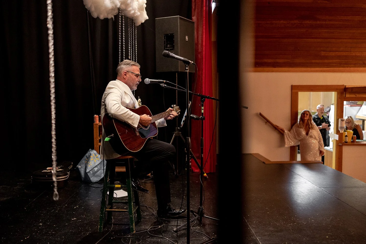 A man in a white blazer playing an acoustic guitar on stage, standing in front of a black curtain with a cloud-like decoration above him, while a few people watch from a distance near a window.