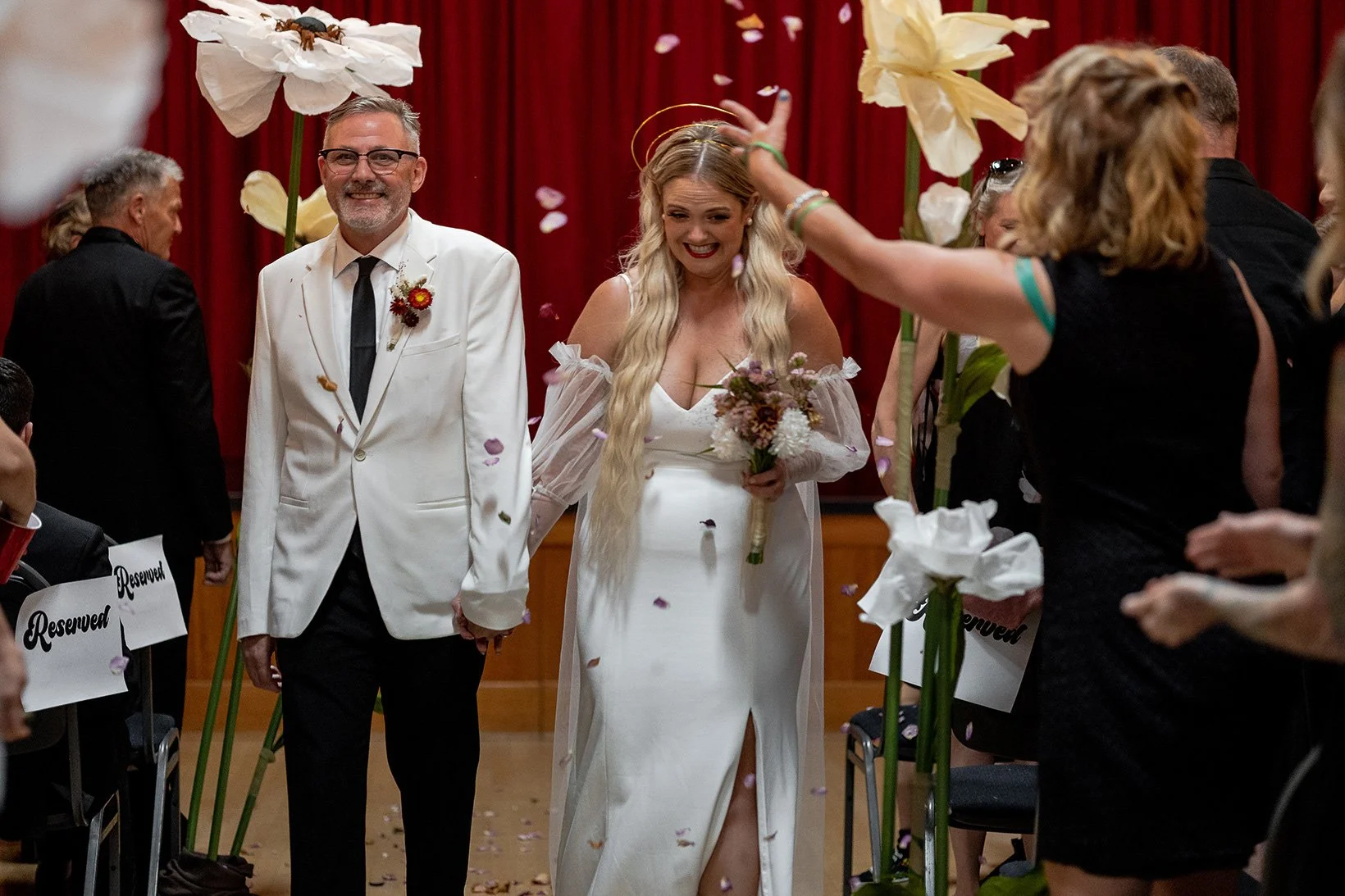 A bride in a white wedding dress holding a bouquet tapping a man in a white suit on the head with her right hand while walking down an aisle, surrounded by guests and falling flower petals, in a decorated indoor setting.