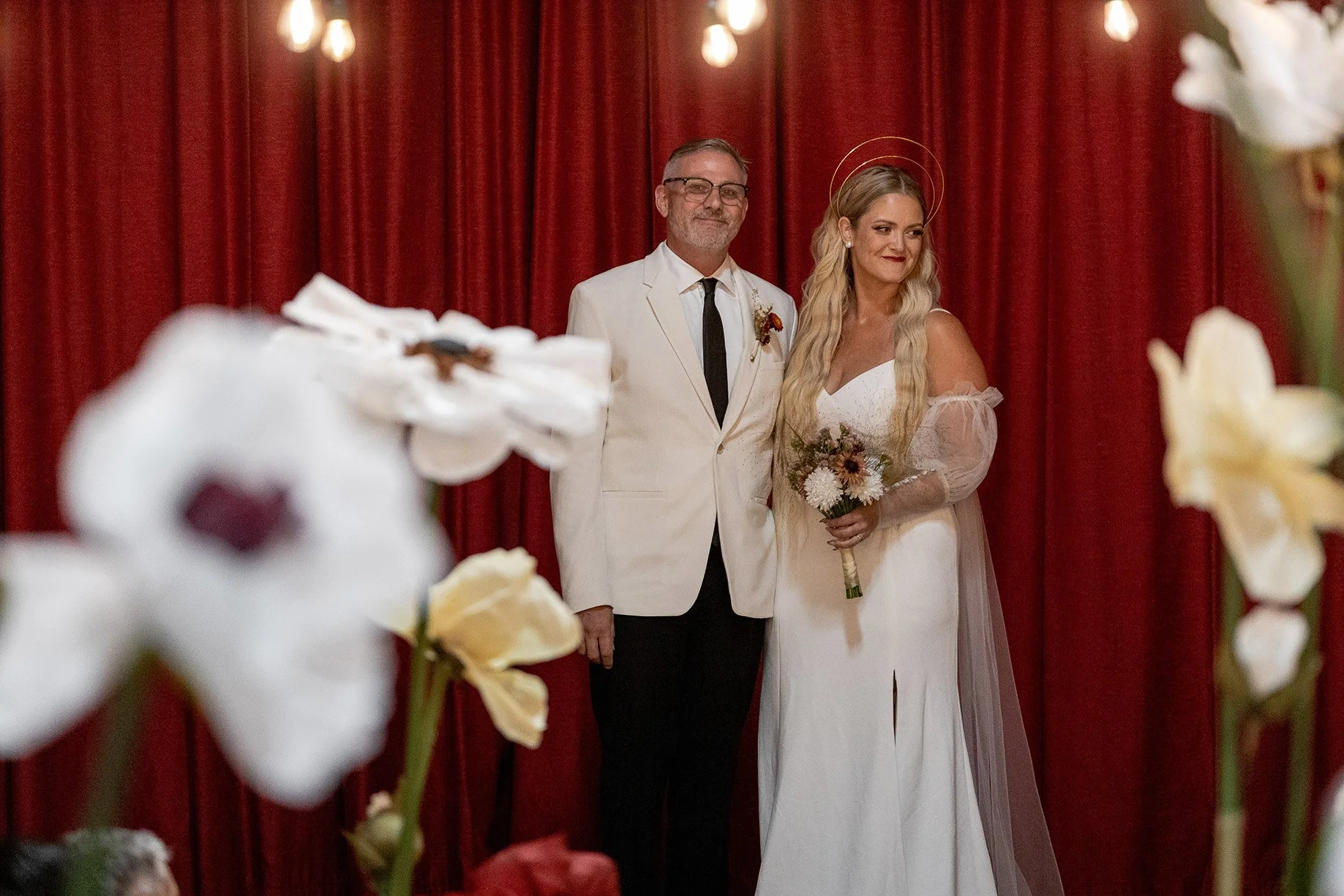 A bride and groom standing together indoors with a red curtain background, surrounded by white flowers. The bride wears a white wedding dress with long sheer sleeves and holds a bouquet, and the groom wears a white suit jacket with black pants and a 