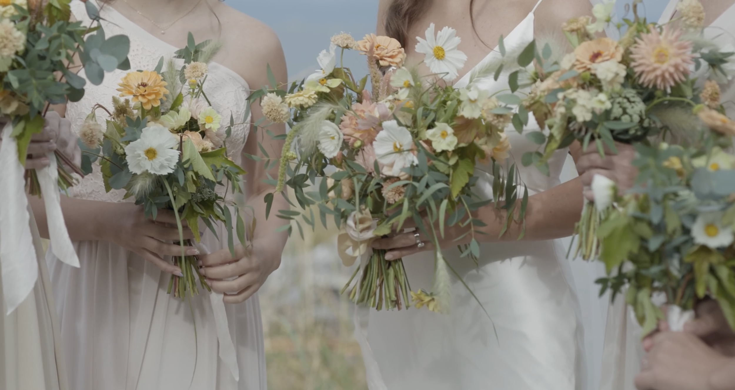 Group of women dressed in light-colored dresses holding flower bouquets during a wedding or outdoor event.
