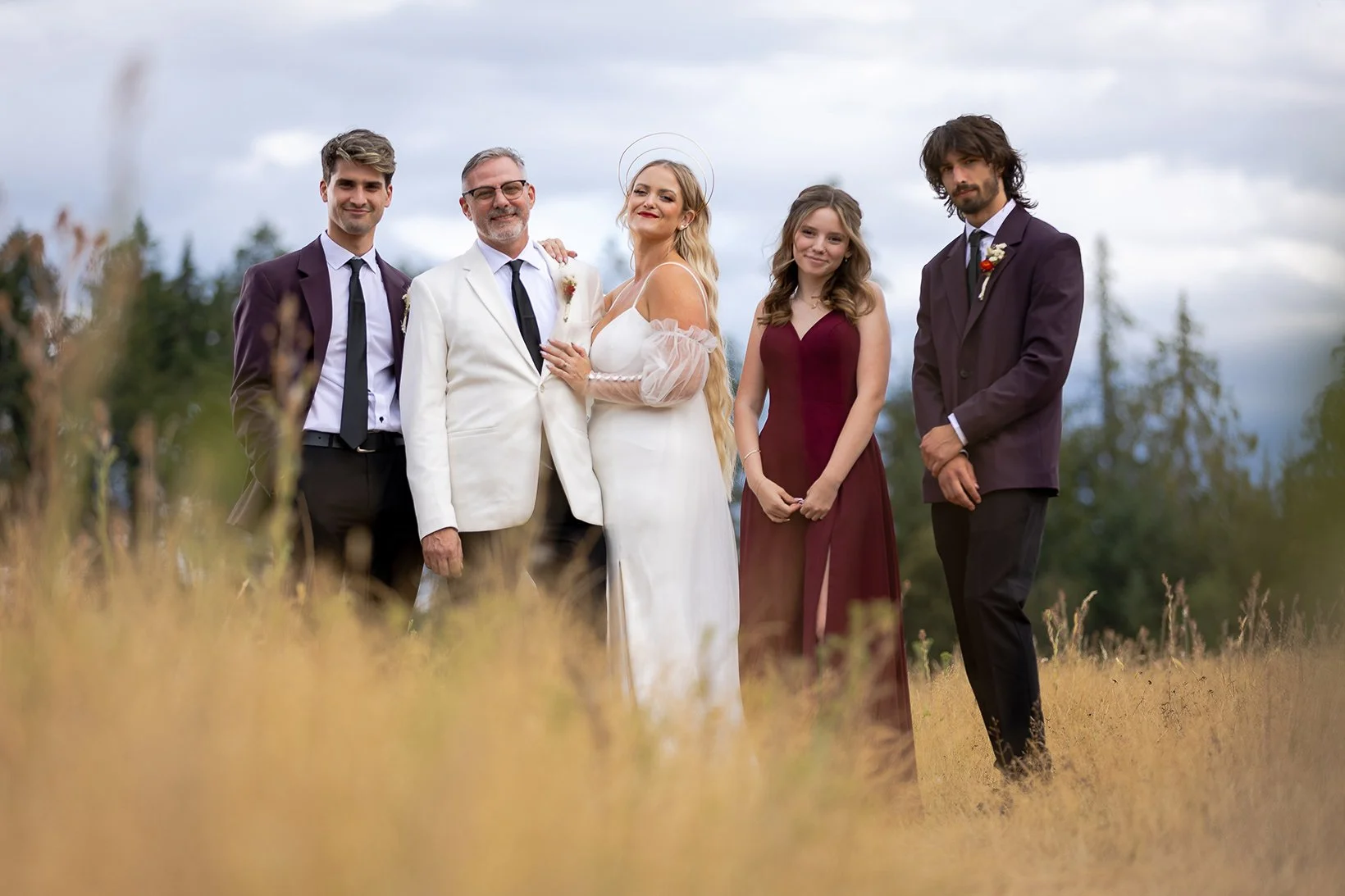 A wedding party outdoors on a cloudy day with six people, including a bride, groom, officiant, and three attendants, standing in a grassy field with trees in the background.