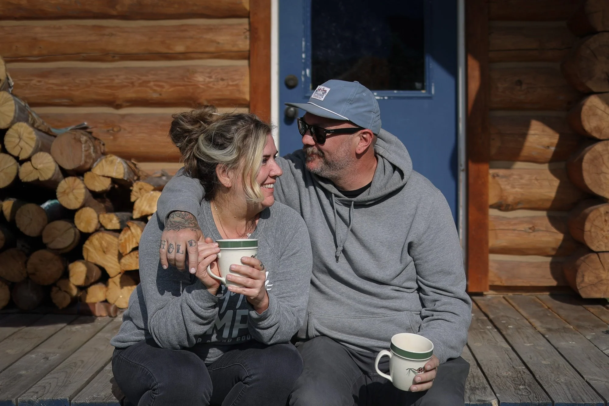 A couple sitting on a wooden porch in front of a log cabin, sharing a moment of affection with coffee mugs in hand.