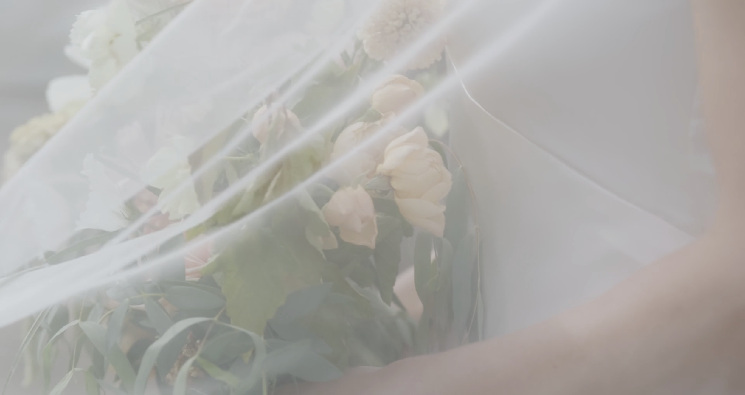 A person holding a bouquet of white and pale pink flowers wrapped in translucent paper.