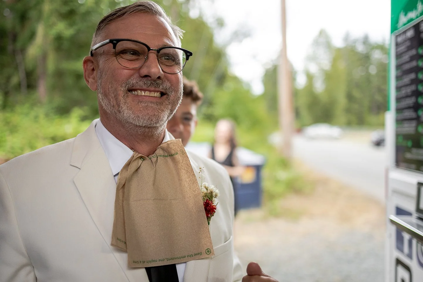 A smiling man with glasses wearing a white suit and a paper bag tie stands outdoors near a menu board, with a blurred background of greenery and people.