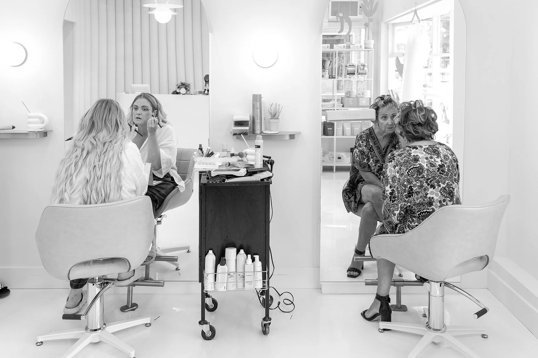 Two women are getting makeup or skincare treatments at a beauty salon, while a third woman is having her makeup or skincare done in the background. The salon has bright lighting, shelves with various products, and a large mirror.