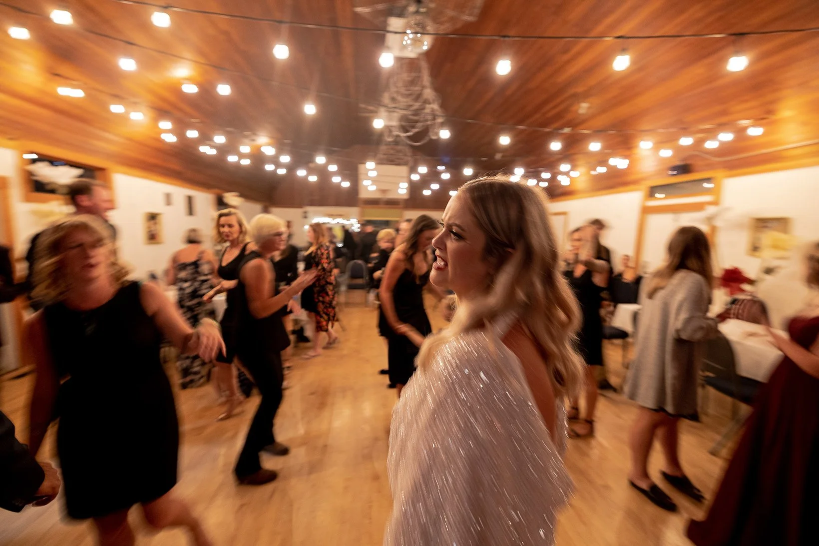 A woman with blonde hair and a glittery dress is dancing and smiling at a party, surrounded by other guests dancing in a warmly lit wooden-floored hall with string lights hanging from the ceiling.