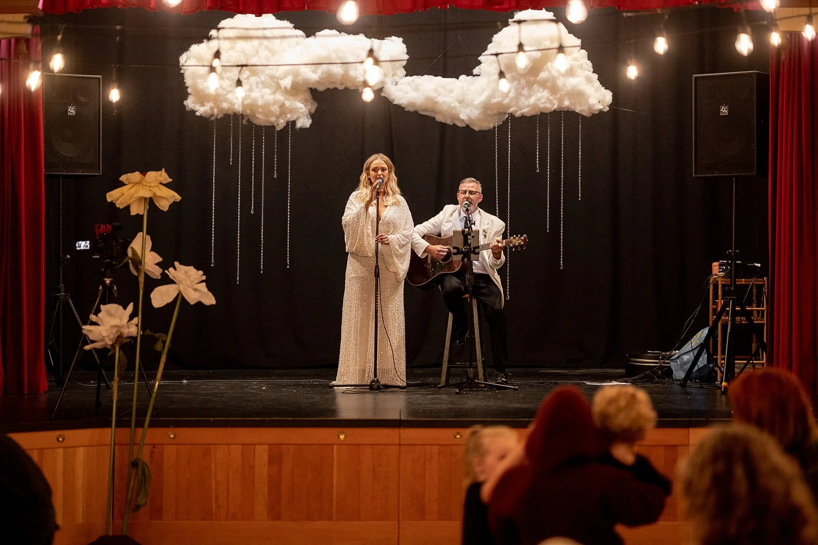 Two performers on stage, one woman singing into a microphone in a shimmering gown and one man playing guitar and singing, with decorative clouds hanging above them.