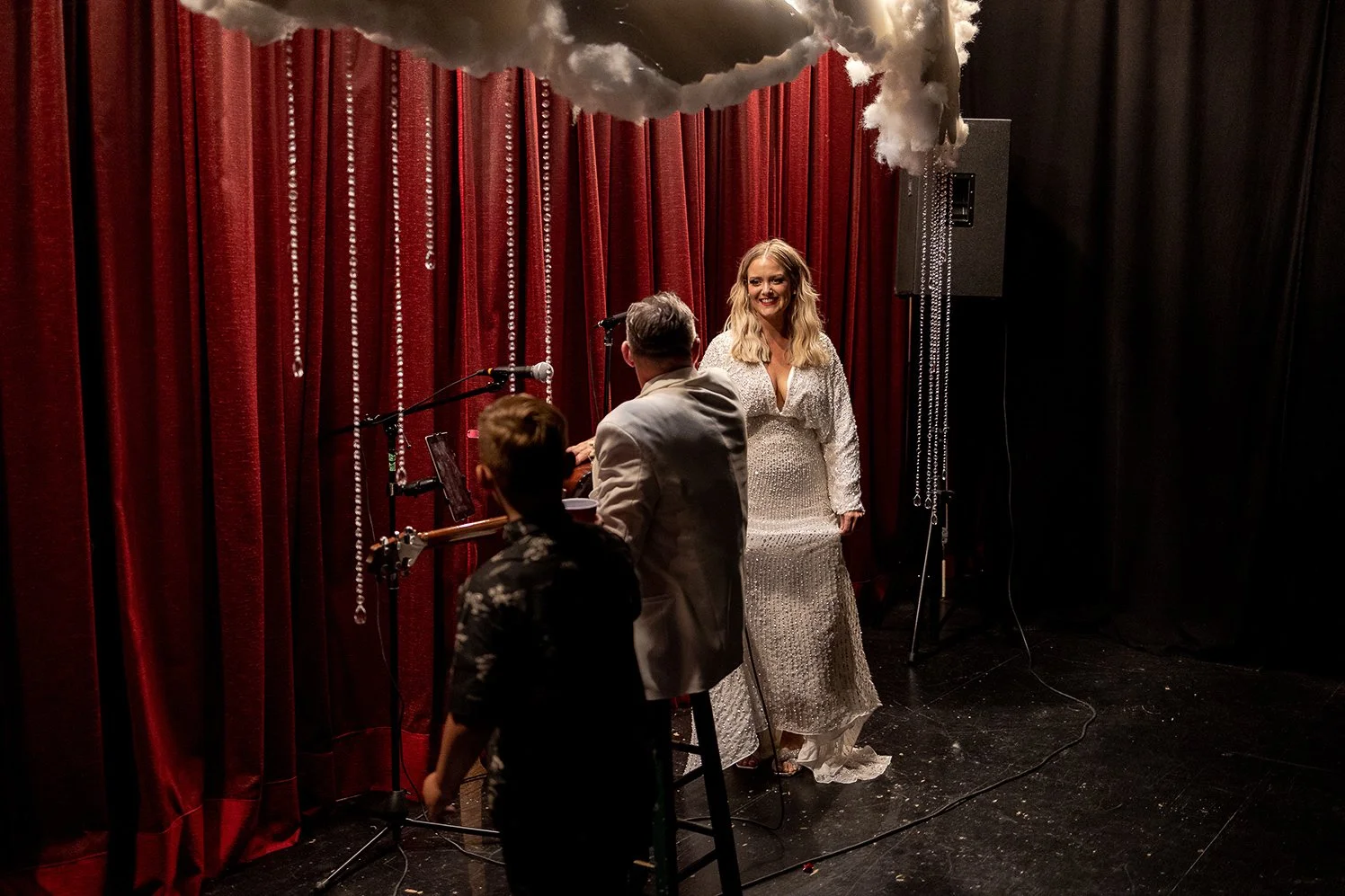 Woman in a sparkly white dress on stage with red curtains and stage fog, while two men, one with gray hair and the other with brown hair, stand nearby, possibly performing or preparing to perform.