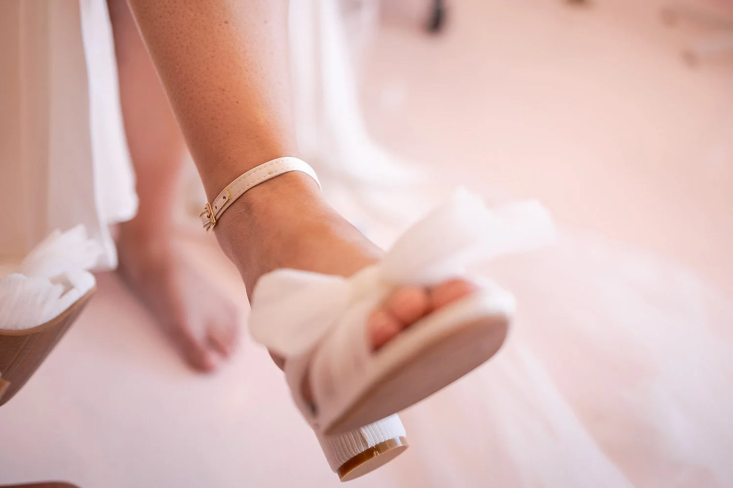 A person wearing a white sandal with a block heel, sitting on a pink surface, with their foot slightly raised. The person is also wearing a white anklet.