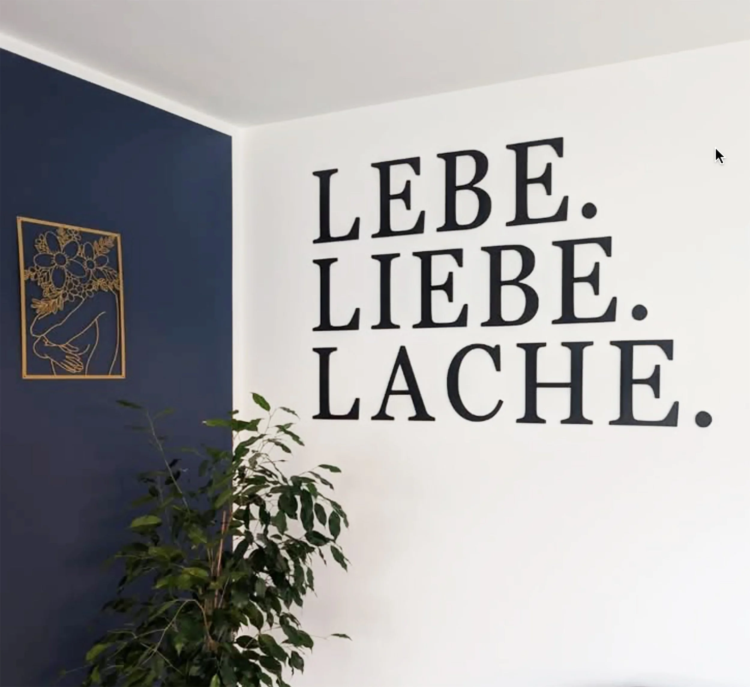 Ein Wandbild mit dem deutschen Text "Lebe. Liebe. Lache." in schwarzer Schrift auf weißem Hintergrund, daneben eine dunkle Wand mit einer goldfarbenen Kunstquelle und eine Topfpflanze.