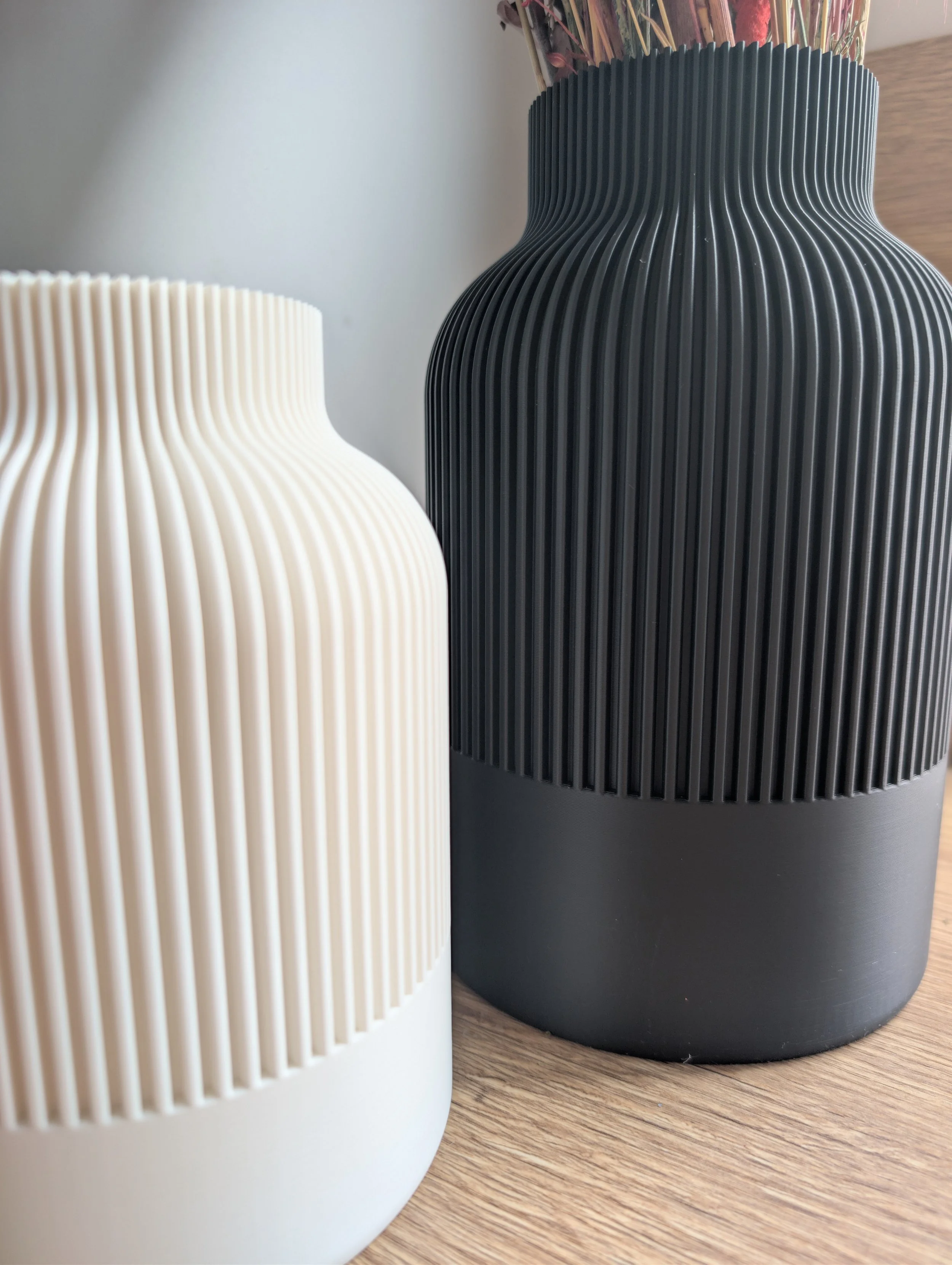 Diana´s Atelier – Zwei dekorative 3D-Druck-Vasen, eine weiße und eine schwarze, mit vertikalen Rillen, auf einem Holztisch. Die Vasen enthalten getrocknete Zweige oder Blumen.