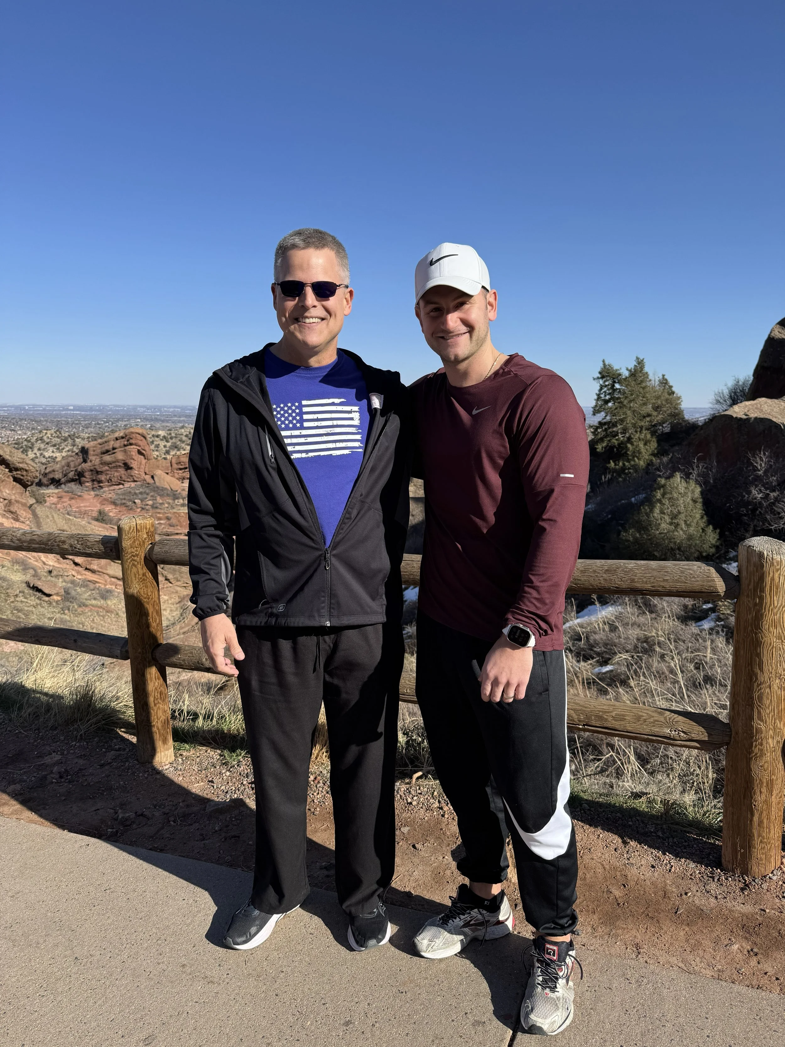 Two men standing outdoors on a trail with desert landscape, rocks, and trees behind them. They are smiling and dressed in athletic clothing, with clear blue sky overhead.
