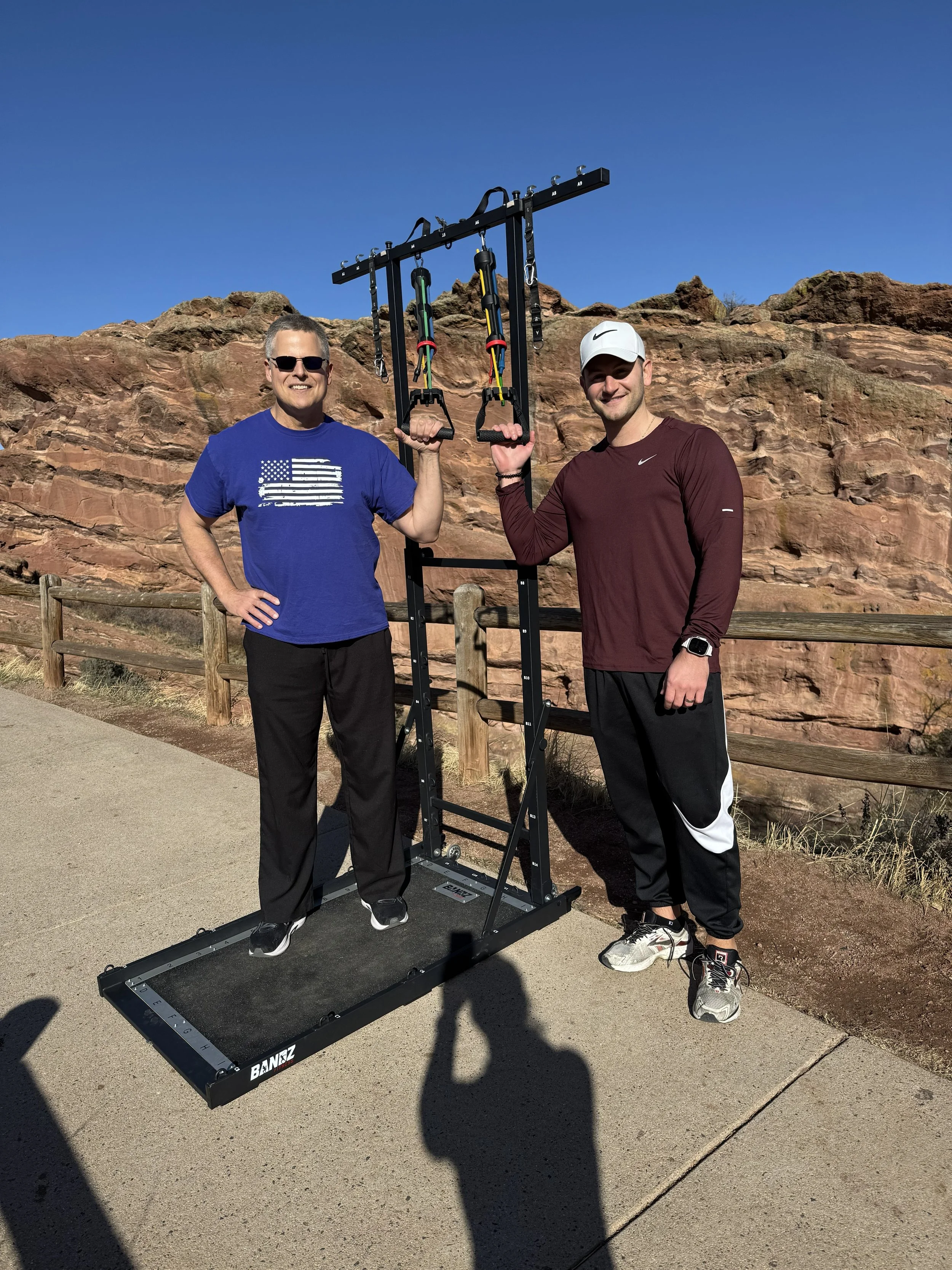Two men standing outdoors in front of a rocky landscape, holding a black fitness machine with resistance bands and handles, smiling at the camera.