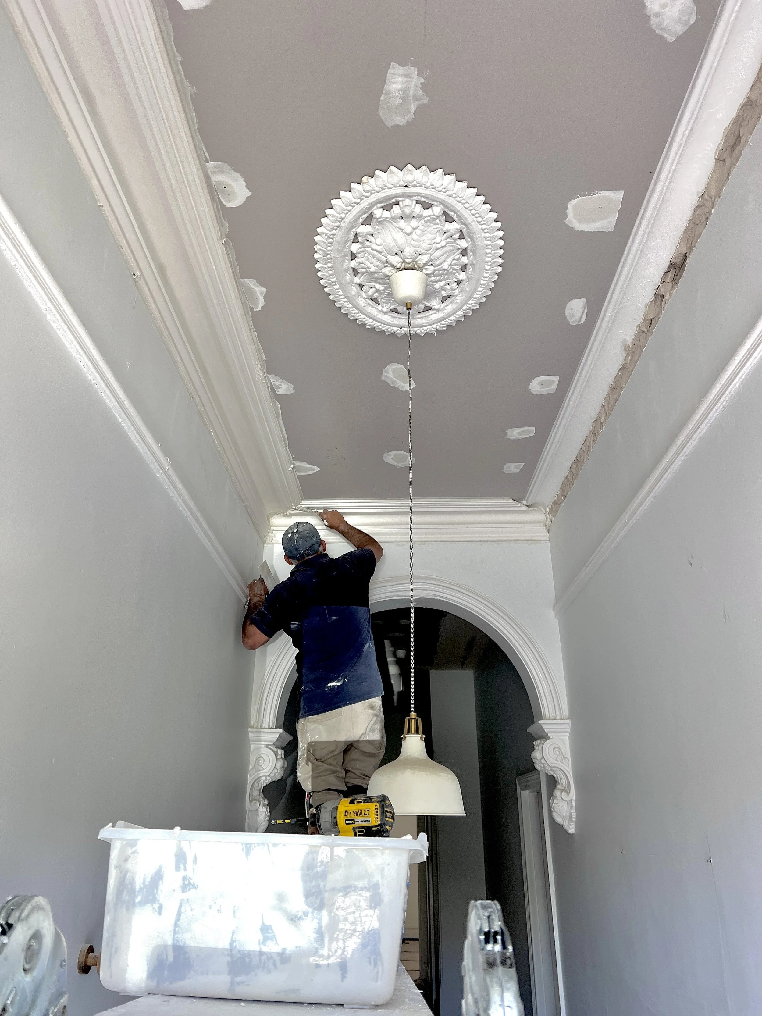 Worker repairing decorative ceiling medallion in a hallway, standing on a bathtub, with a drywall hole patching process visible on the ceiling.