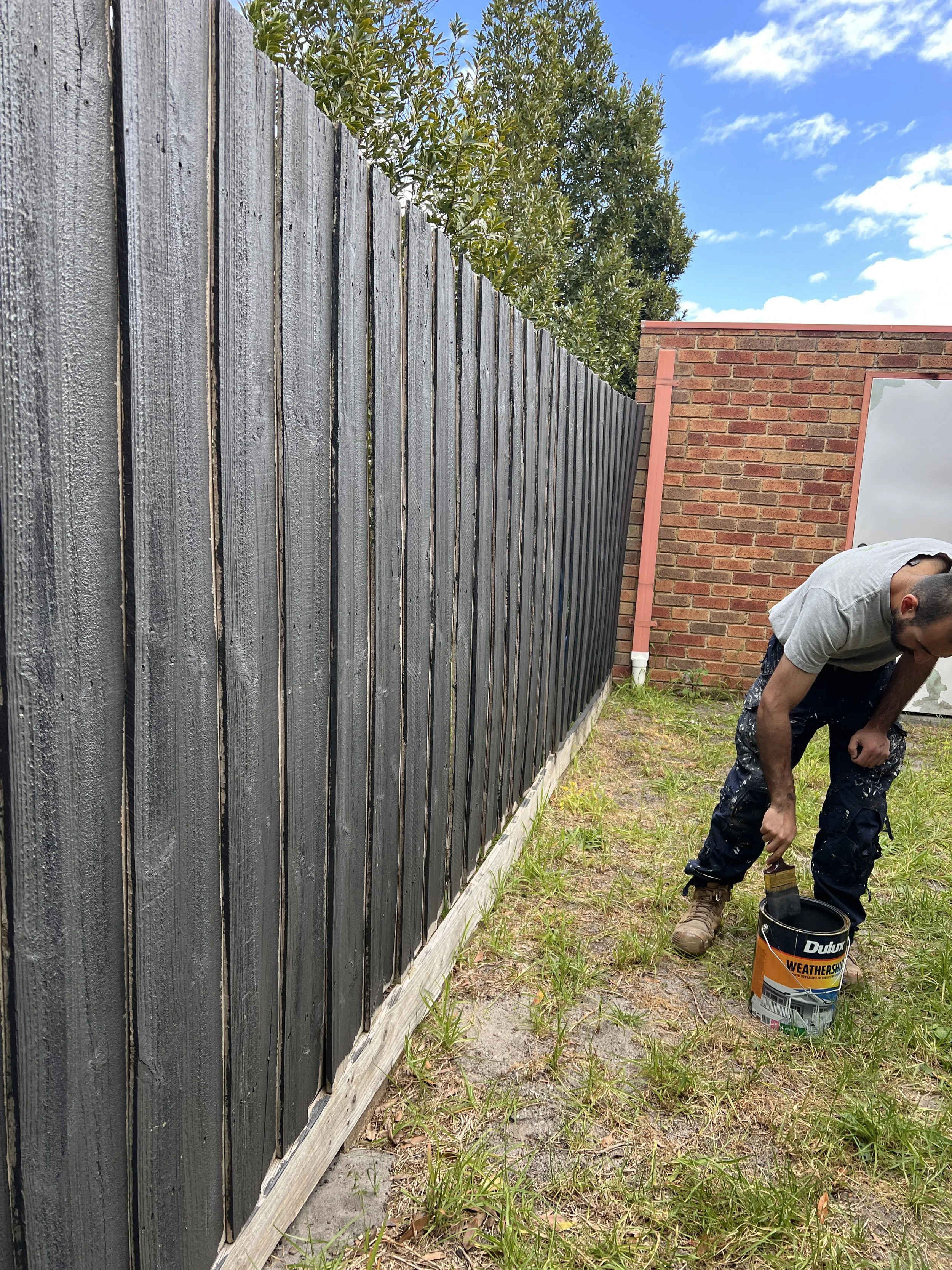 A man painting a wooden fence with black weatherproof paint in a backyard, near a brick house and some green trees.