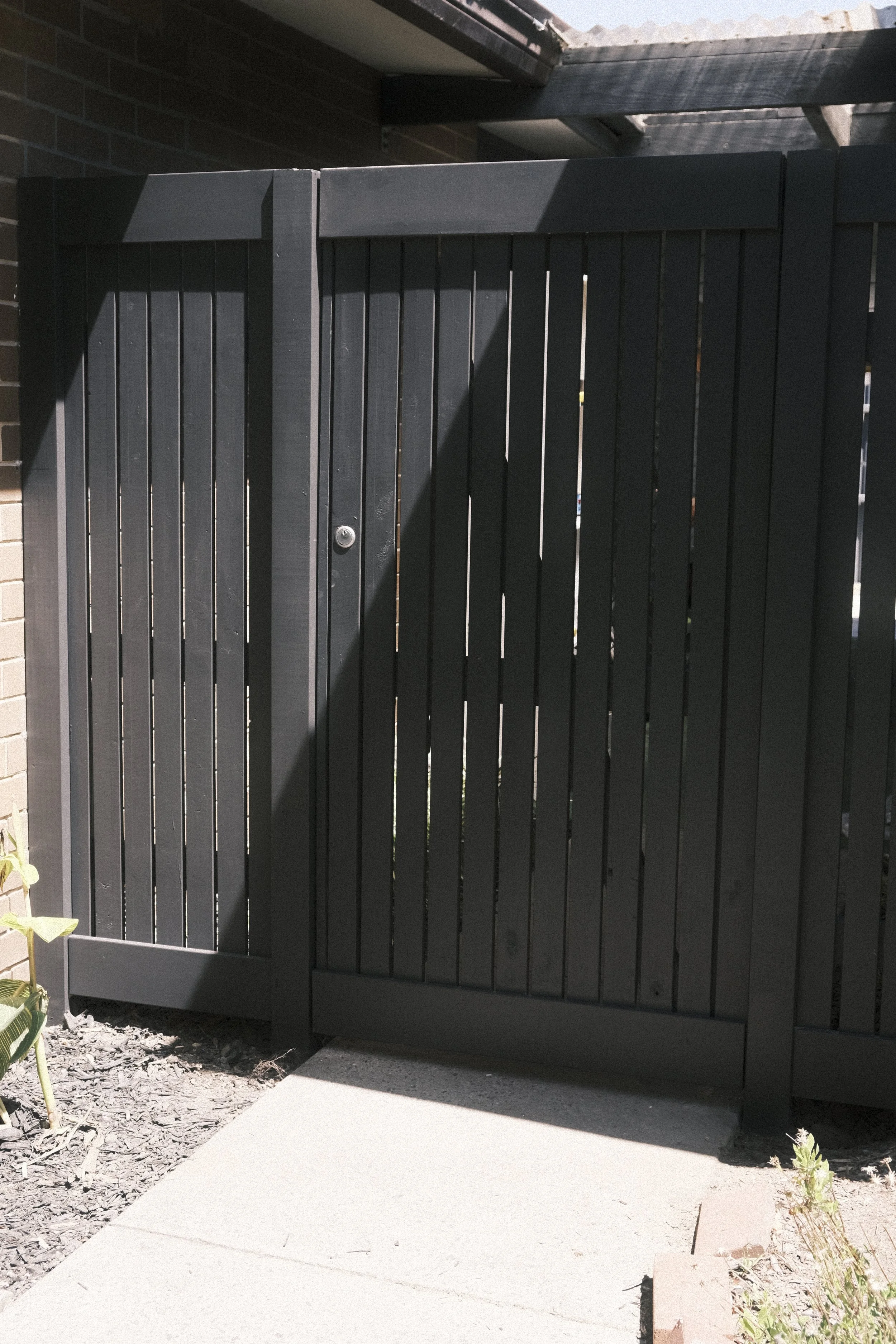 A black wooden gate with vertical slats, installed on a concrete sidewalk, partially shaded. The gate is adjacent to a brick wall and has a small round lock or handle near the center.