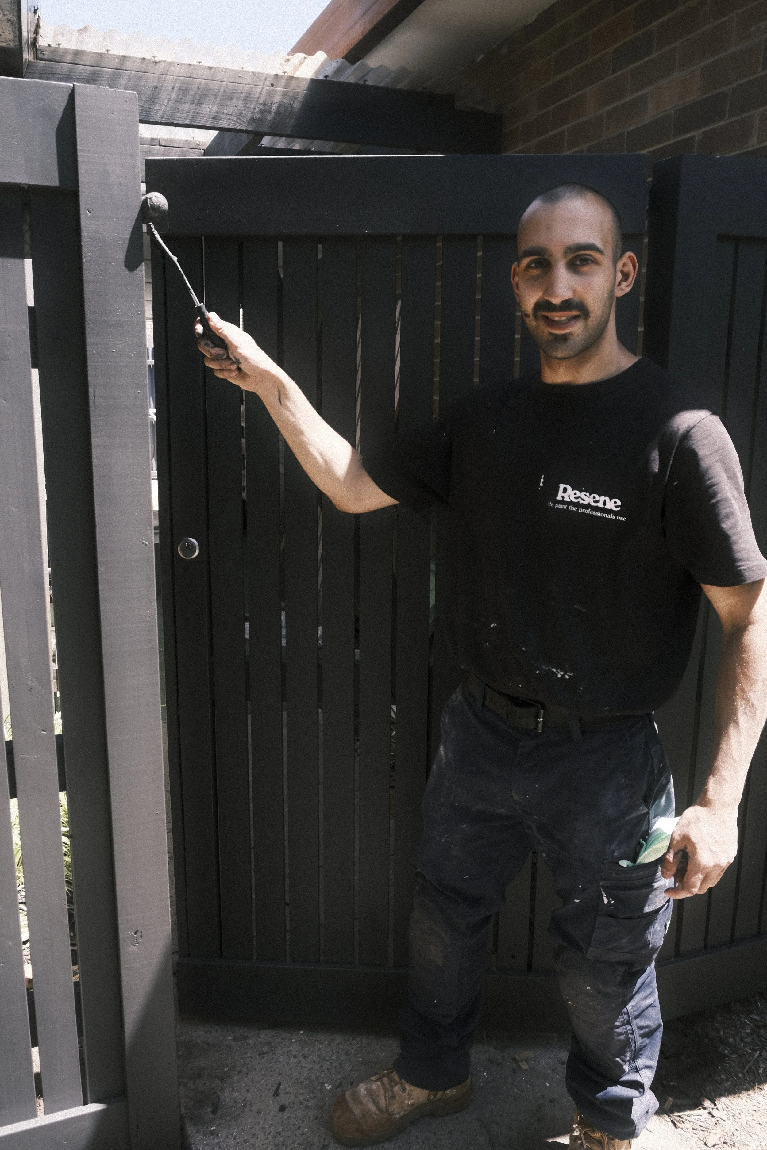 A man is painting a black wooden fence outside in the daytime. He is smiling, wearing work clothes with paint stains.