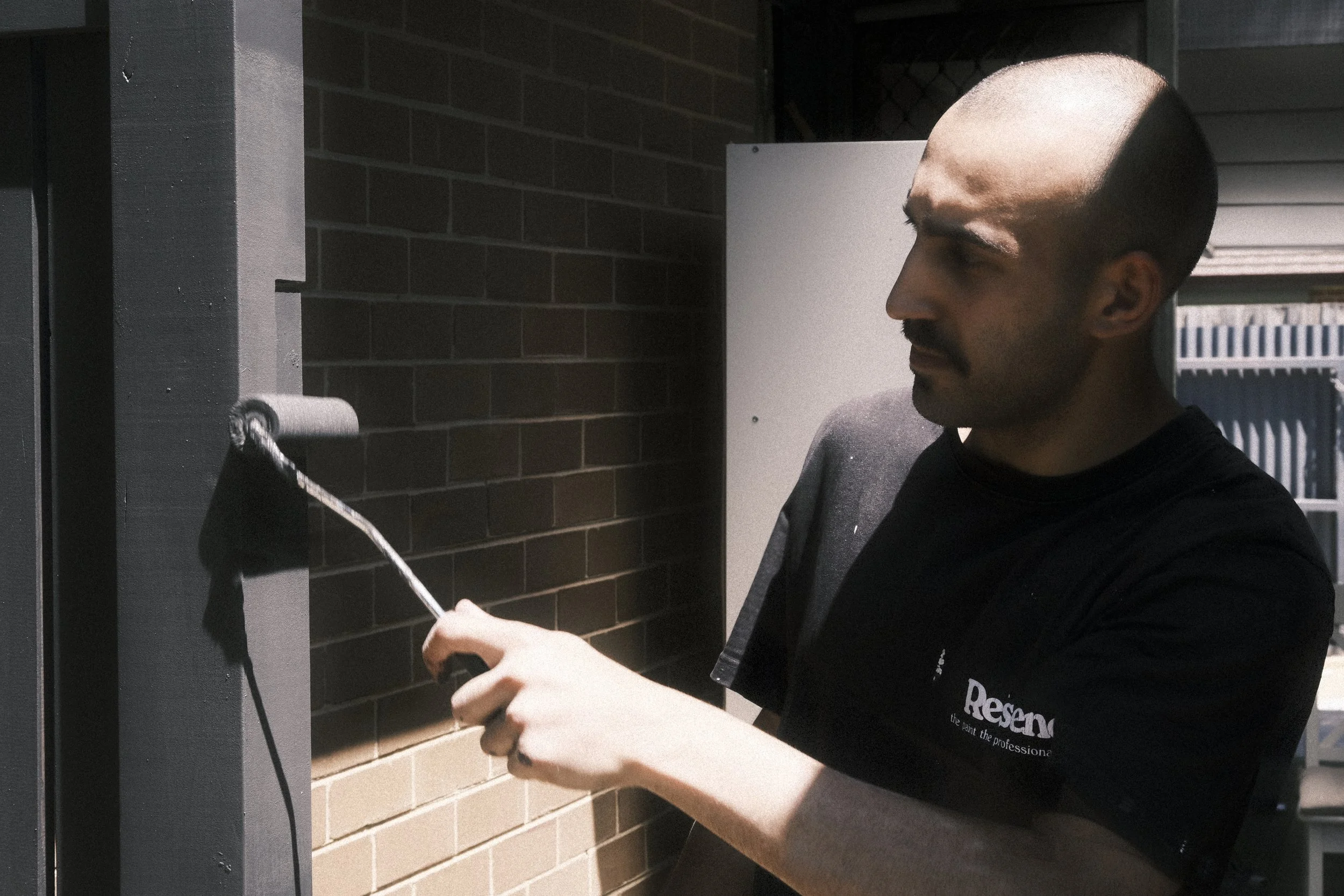 A man painting a wall with a paint roller, standing outside near a brick wall, wearing a black shirt.