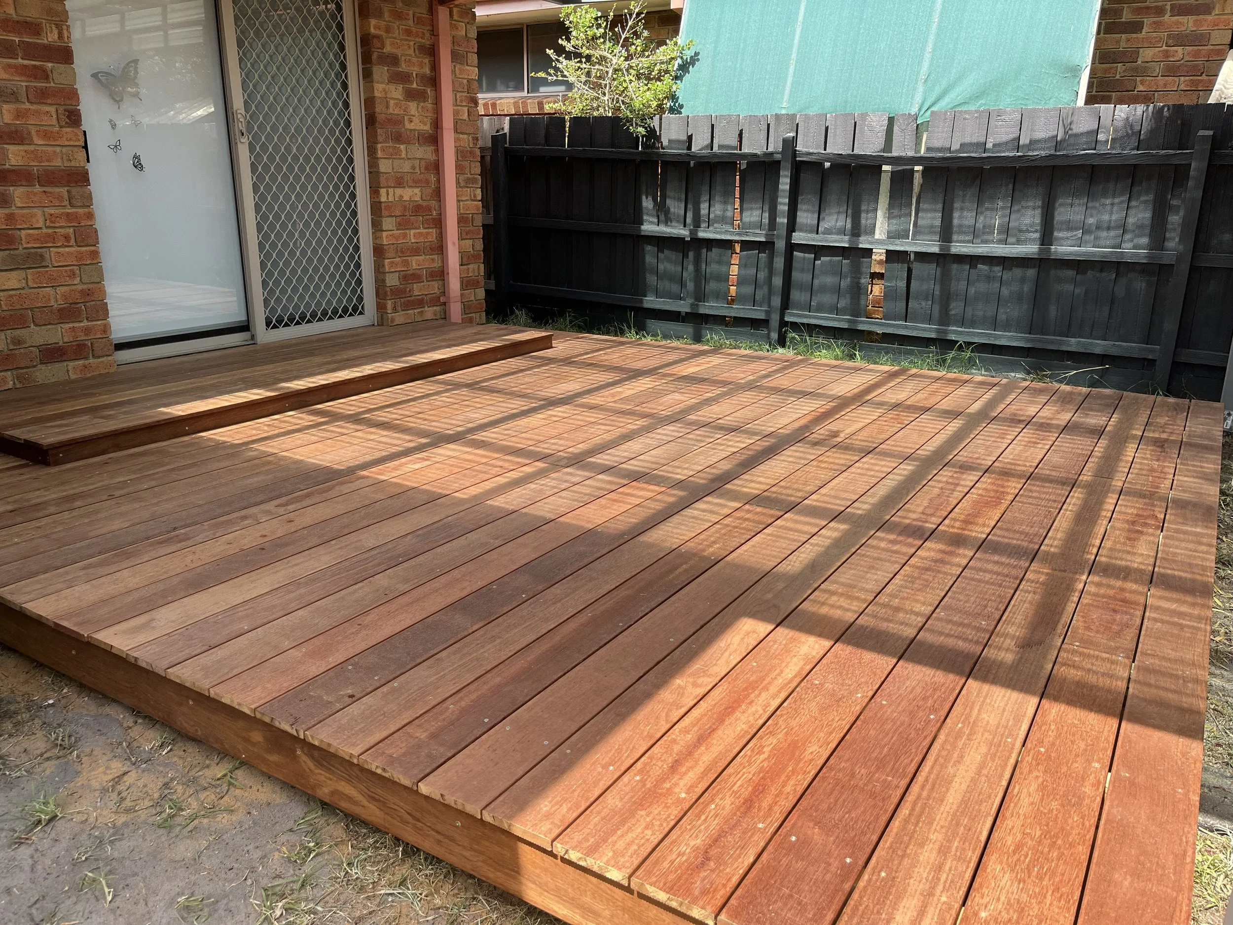 Newly constructed wooden deck outside a house with a sliding glass door and a mesh screen, surrounded by a black wooden fence and some greenery.