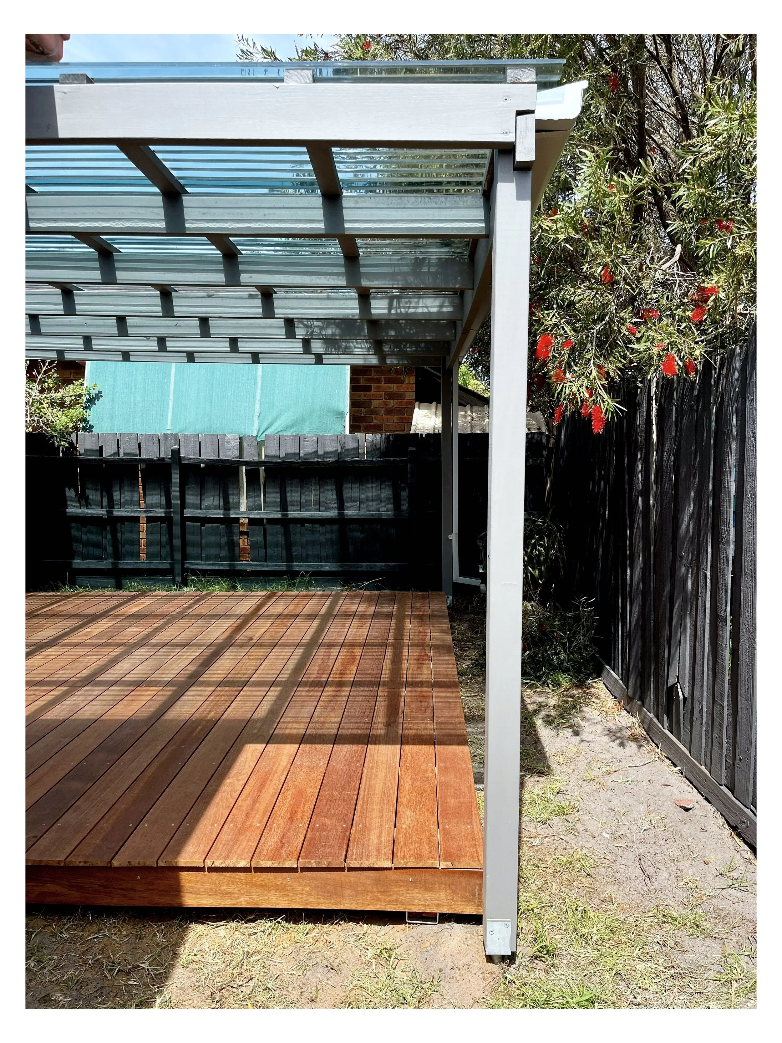 A backyard patio with a wooden deck, a black fence, and a metal pergola with a translucent roof, surrounded by trees with red flowers.