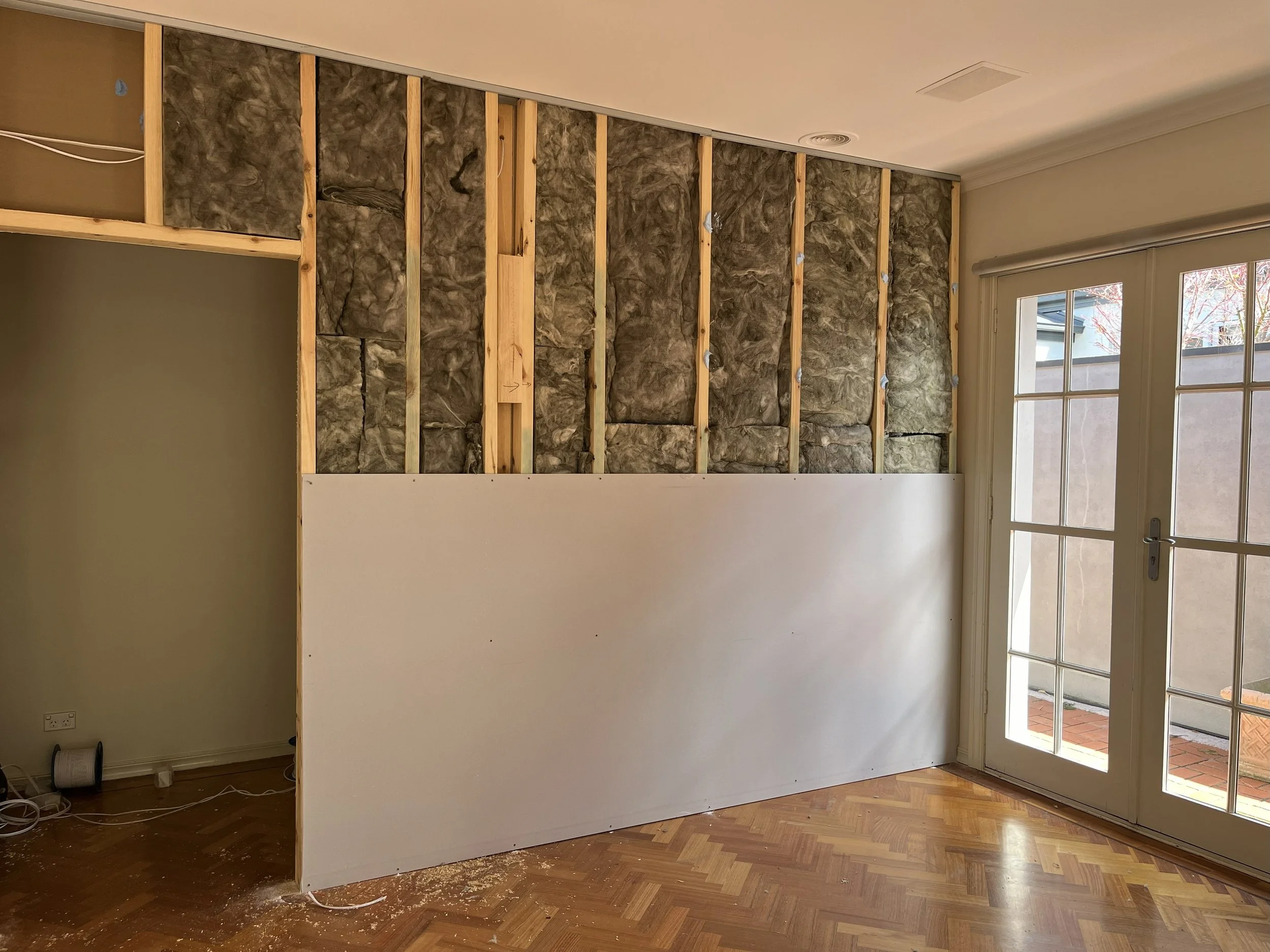 Room with wall under construction, insulation and drywall partially installed, large glass door to outside, hardwood floor.