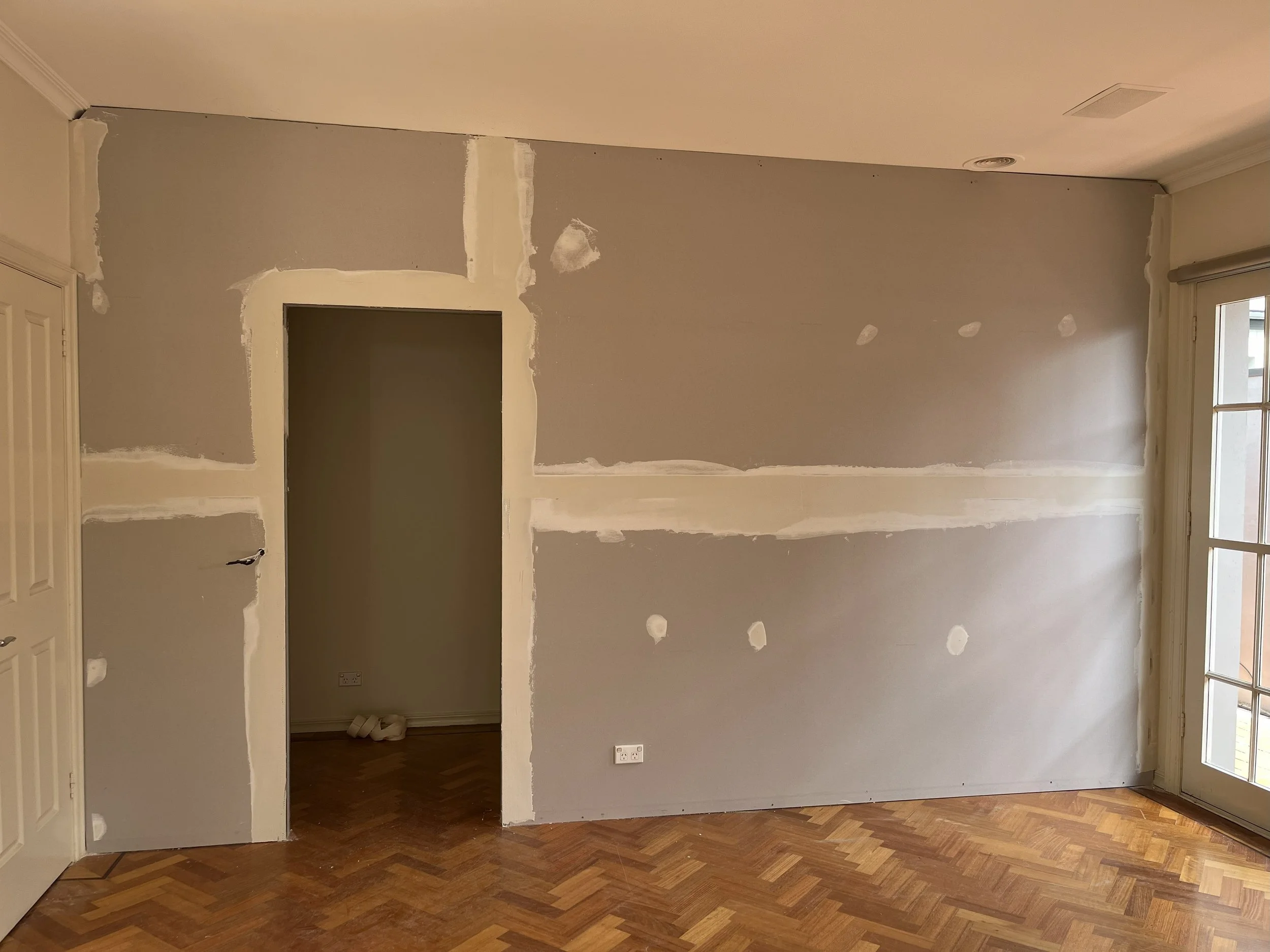 Room with drywall in progress, patches and sanding marks visible, wooden flooring, window, and door opening