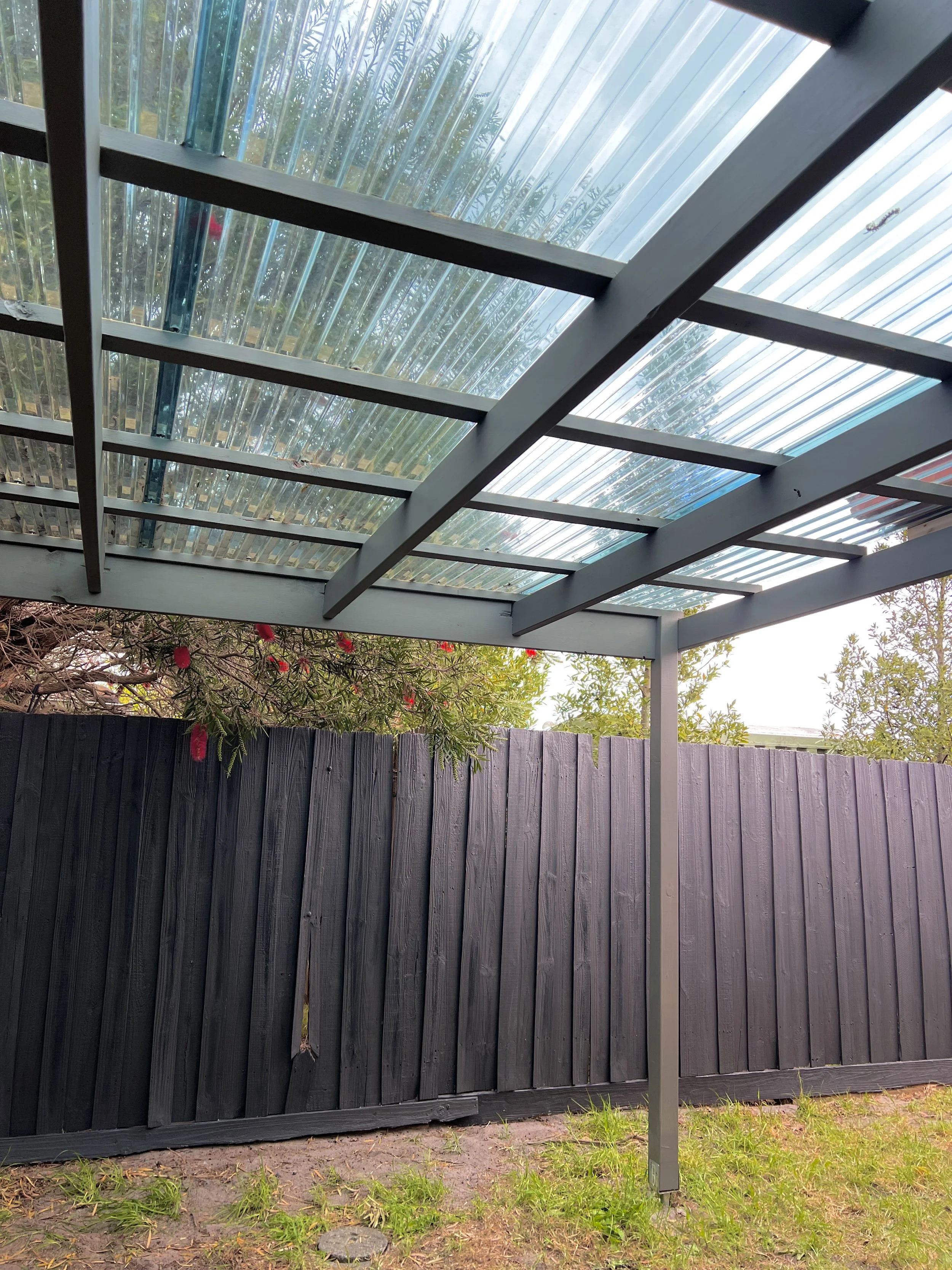 View of a backyard patio with a transparent corrugated plastic roof supported by timber  beams, a black wooden fence, and some plants with red flowers visible behind the fence.