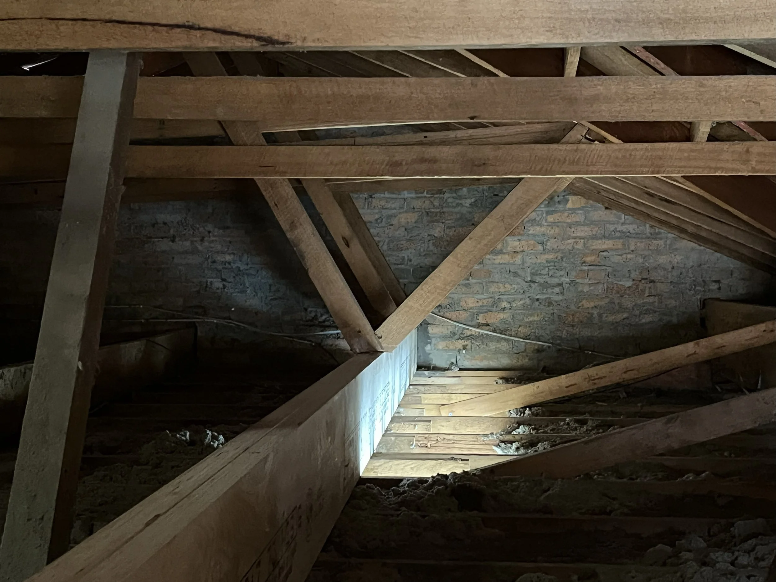 View looking through an unfinished attic or roof space with exposed wooden beams, rafters, and a brick wall in the background.