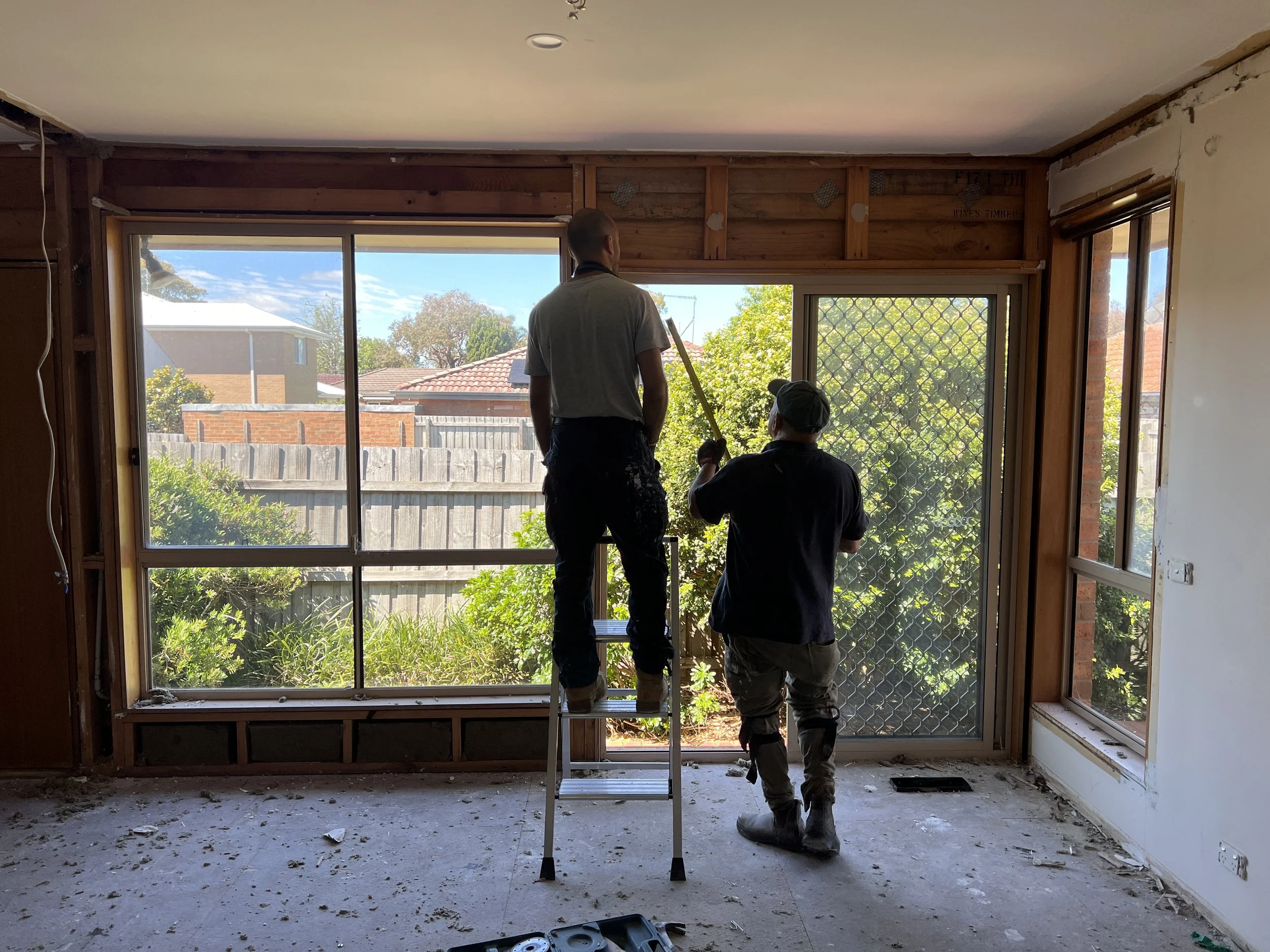 Two workers are installing a large window in a house, with one standing on a ladder and the other holding tools, during daylight.