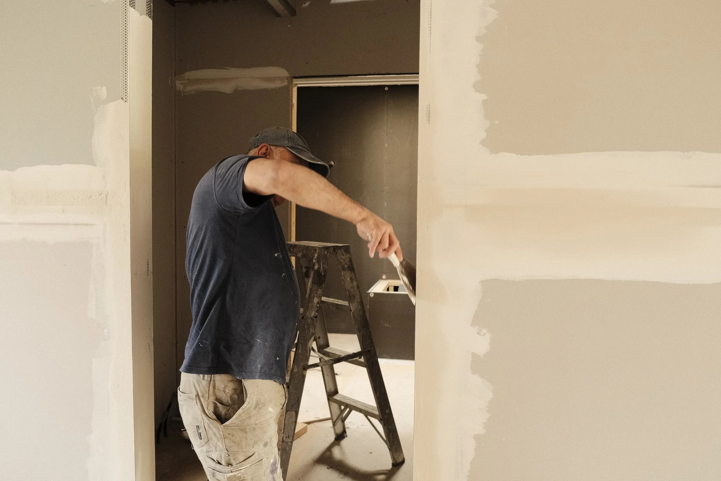 A man in a baseball cap painting drywall in a room under construction, with a stepladder nearby.