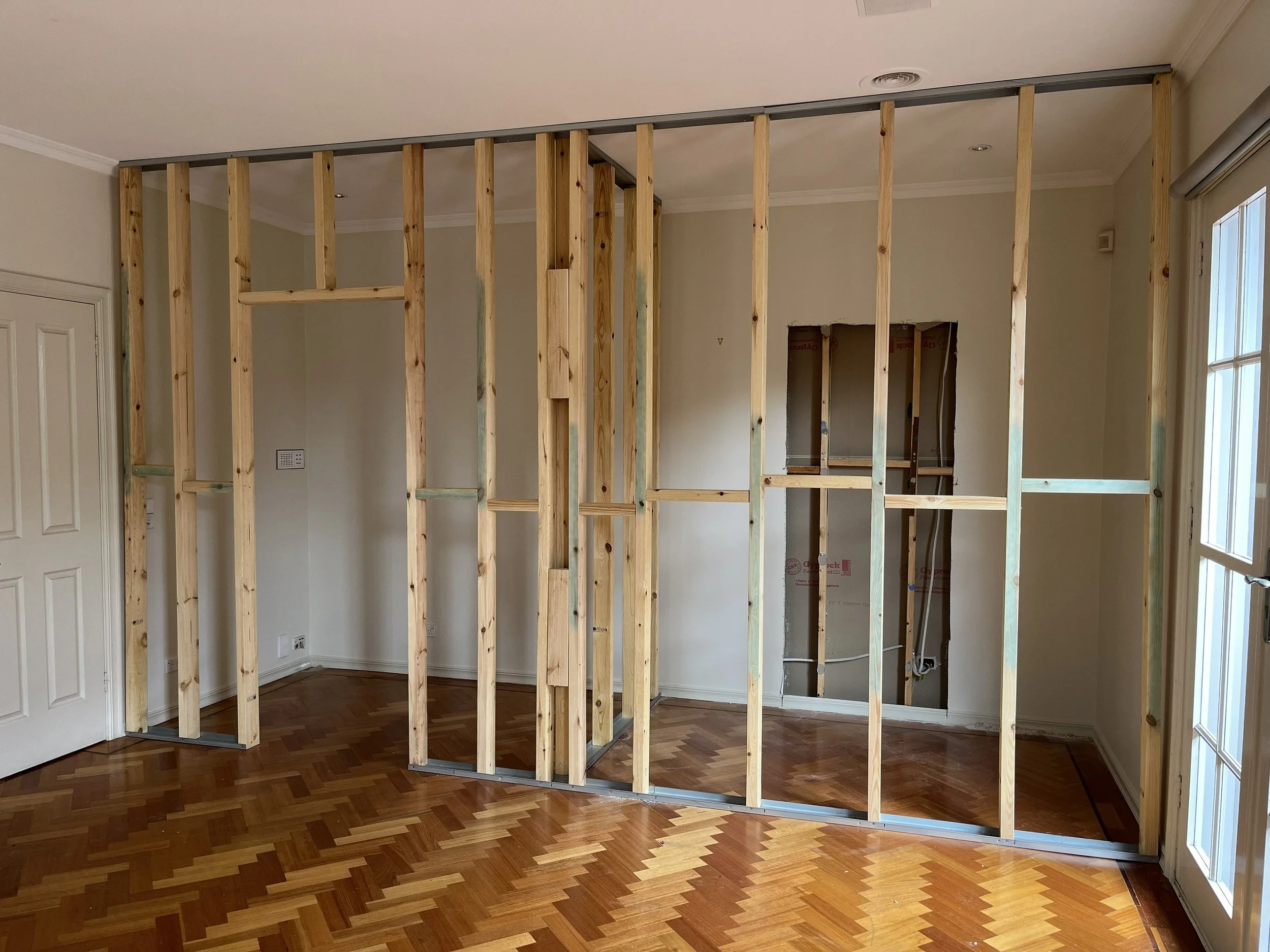 Interior of a room with a wooden stud wall under construction, with no drywall, and a small open doorway in the wall.