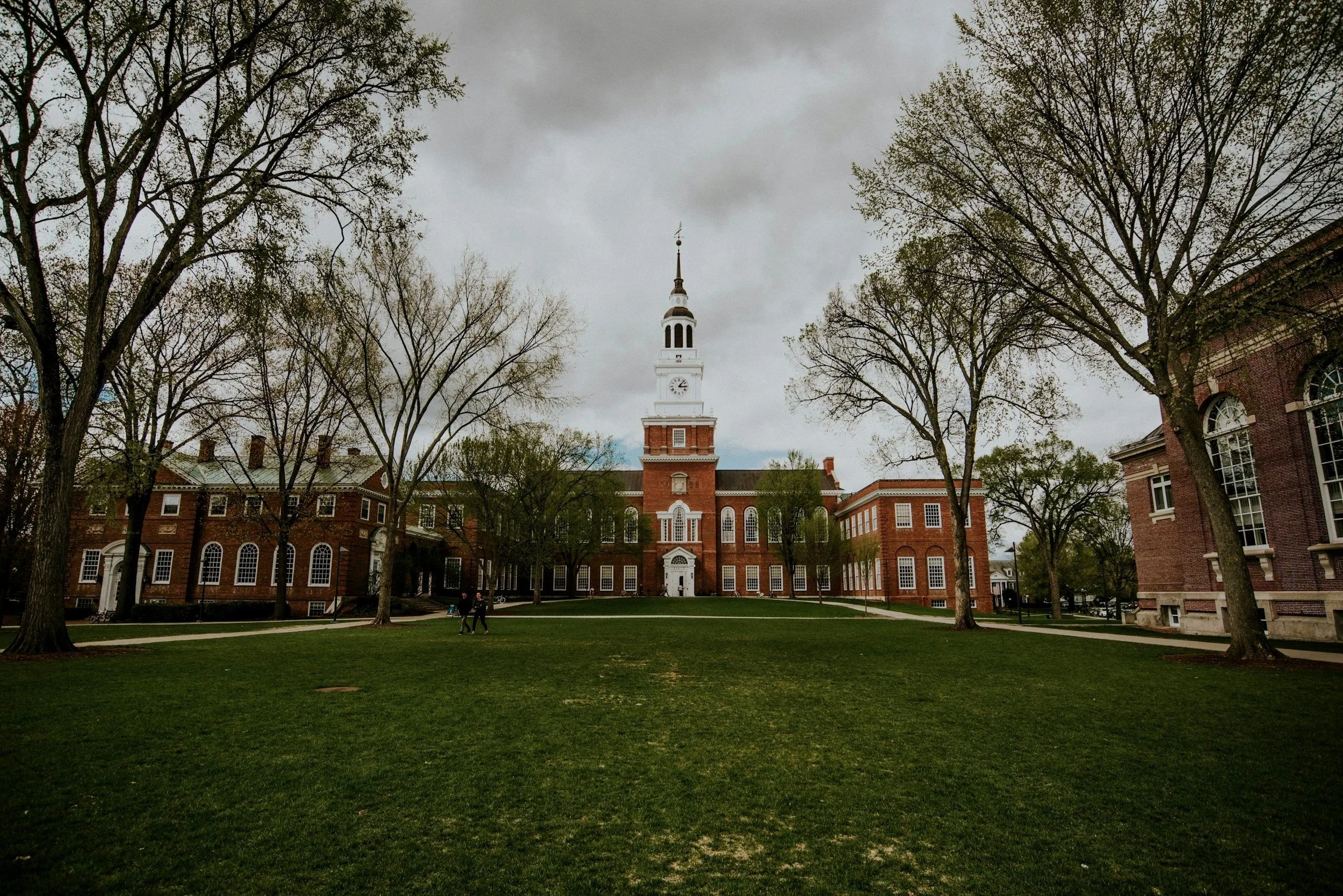 Historic red brick building with a tall white clock tower, surrounded by leafless trees and a grassy area, under a cloudy sky.